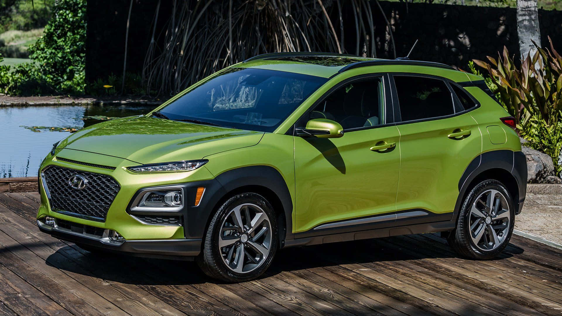 Hyundai Kona 桌布 壁紙 — 一輛充滿活力的萊姆綠色現代 Kona SUV 停泊在寧靜池塘旁的木甲板上，周圍有茂密的熱帶植物。
