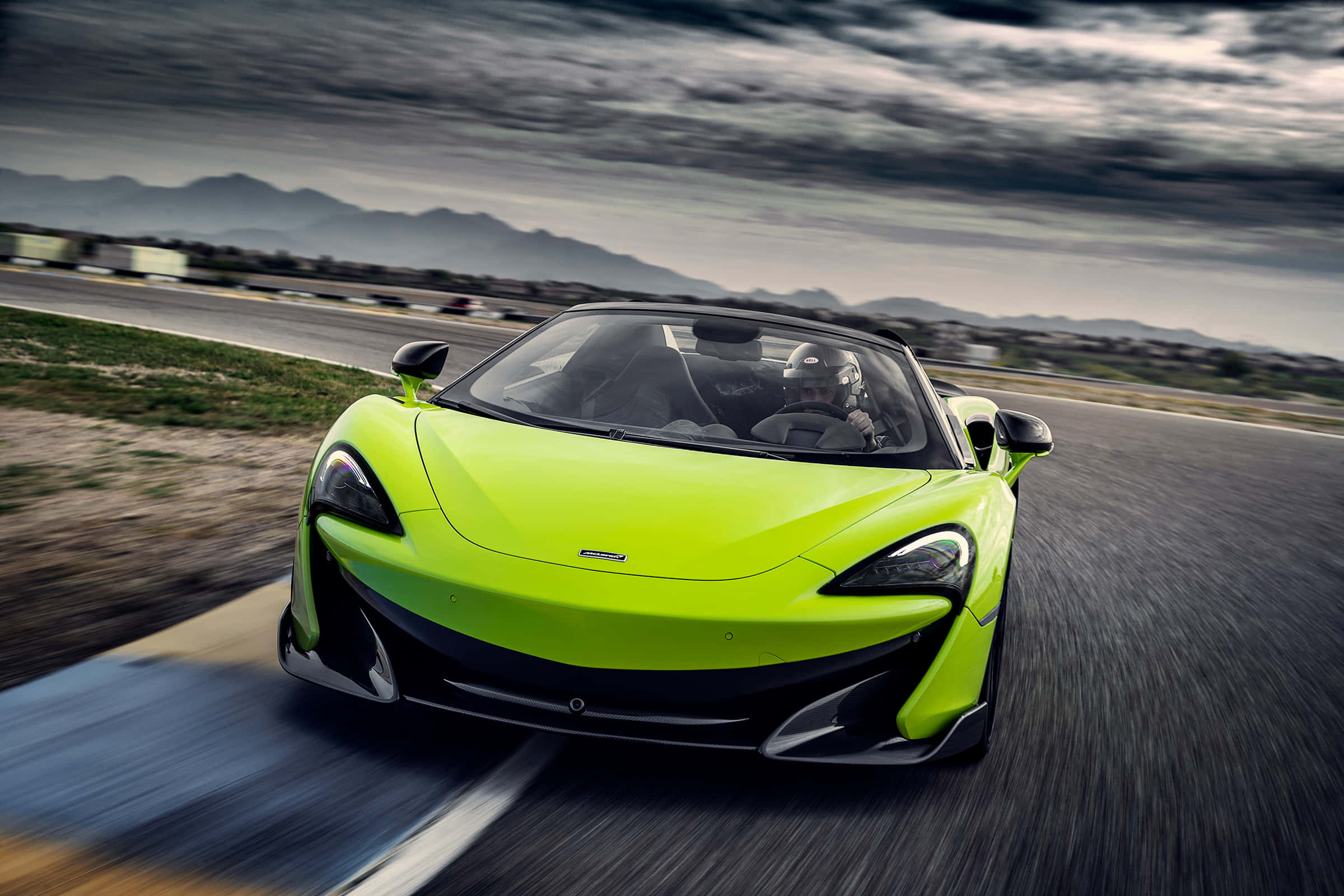 Mclaren 600lt Spider 桌布 壁紙 — 一輛充滿活力的萊姆綠色McLaren 570S敞篷跑車在戲劇性的多雲天空下，以模糊的動態在賽道上奔馳。