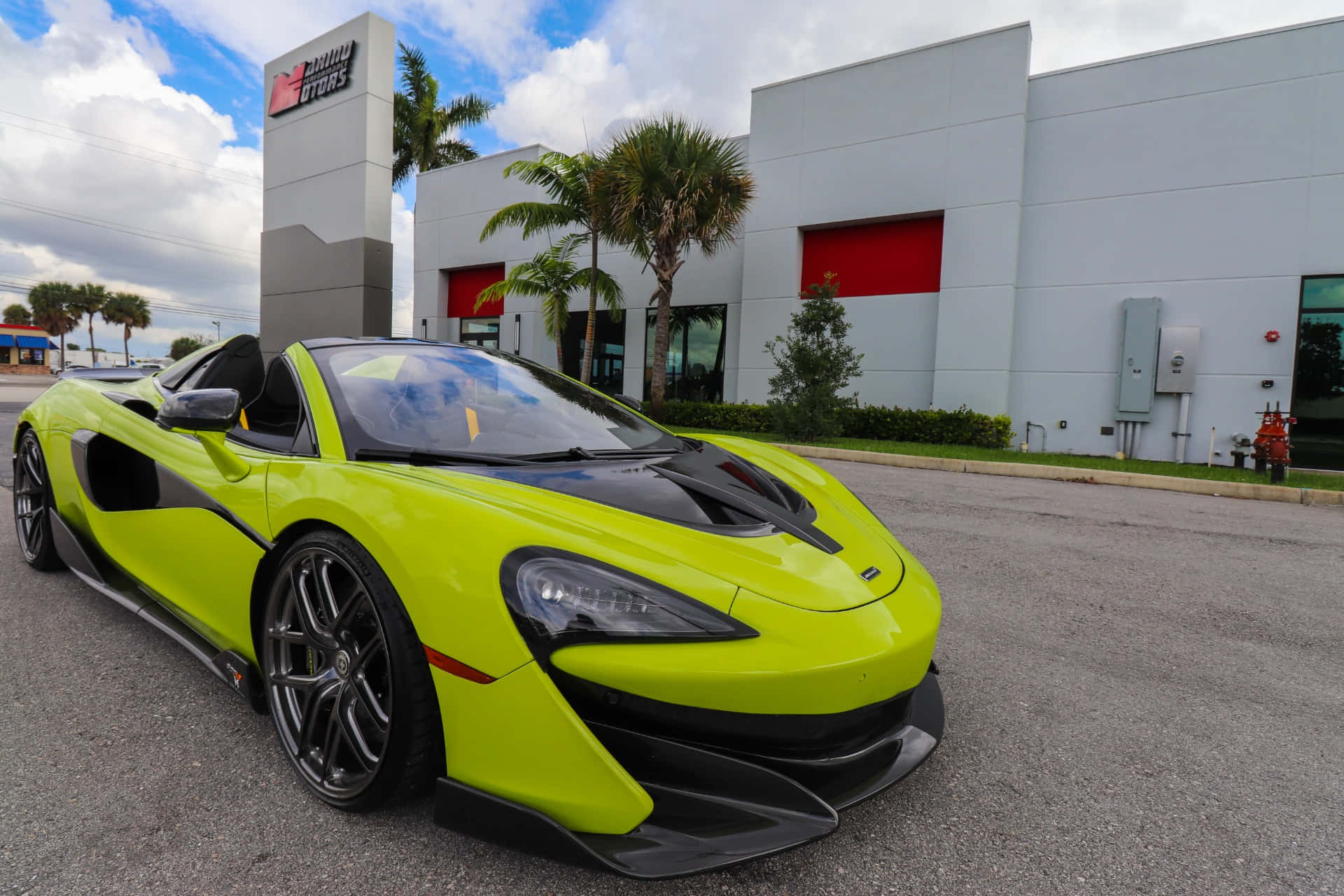 Mclaren 600lt Spider 桌布 壁紙 — 一輛充滿活力的青檸綠色McLaren跑車停放在現代化經銷商外，有棕櫚樹和部分多雲的天空。