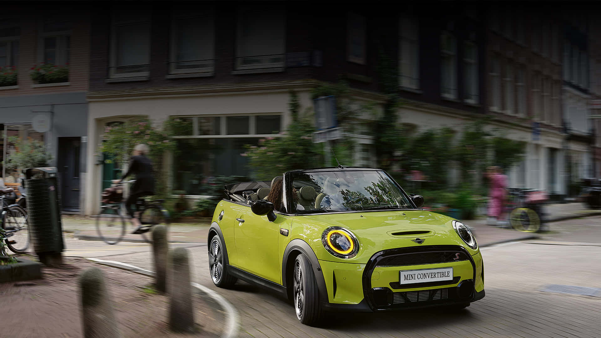 Mini Cooper Convertible 桌布 壁紙 — 一輛充滿活力的萊姆綠色MINI Convertible在歐洲鵝卵石街道上行駛，背景有模糊的自行車騎士和建築物。