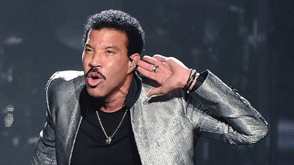 superstar 桌布 壁紙 — Lionel Richie 在舞台上表演，身穿閃亮銀色外套，在舞台燈光下唱歌時手放在耳邊。