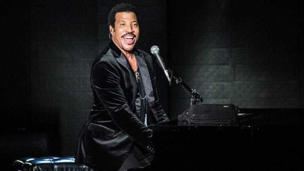 concert 桌布 壁紙 — Lionel Richie 在黑暗的音樂會場景中，在舞台燈光下彈奏鋼琴，露出燦爛的笑容。