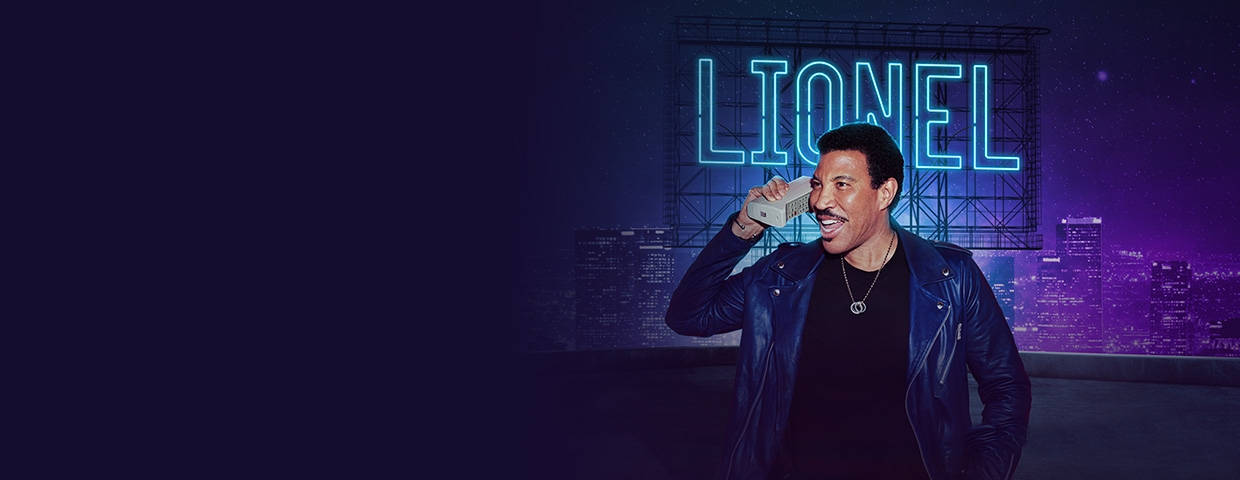 concert 桌布 壁紙 — Lionel Richie 微笑著，手持電話，背景是夜晚霓虹燈點亮的城市景觀，身後有發光的「LIONEL」標誌。