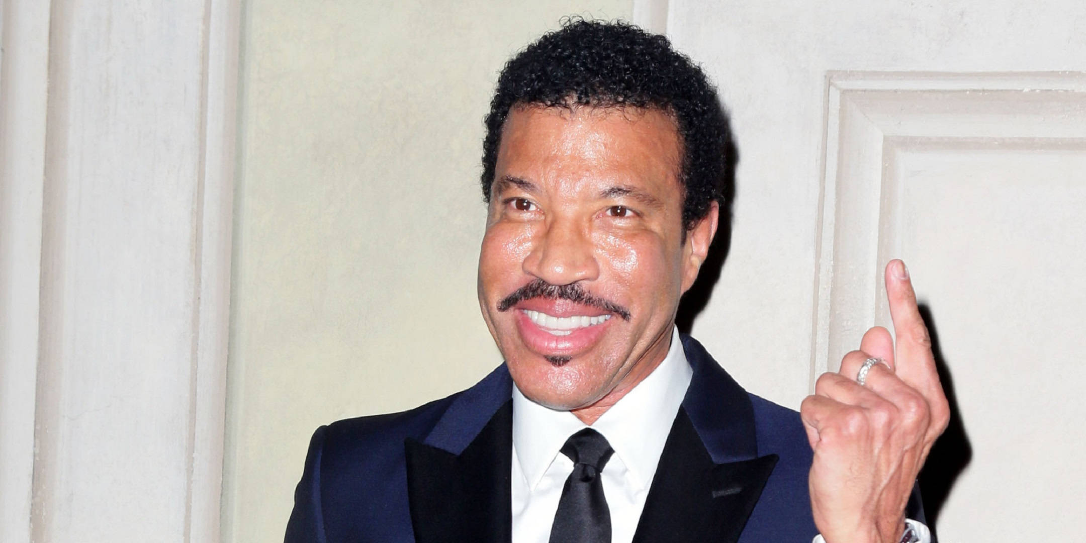 Celebrity 桌布 壁紙 — Lionel Richie 身穿深色西裝，在淺色鑲板牆前溫暖微笑，右手向上指。