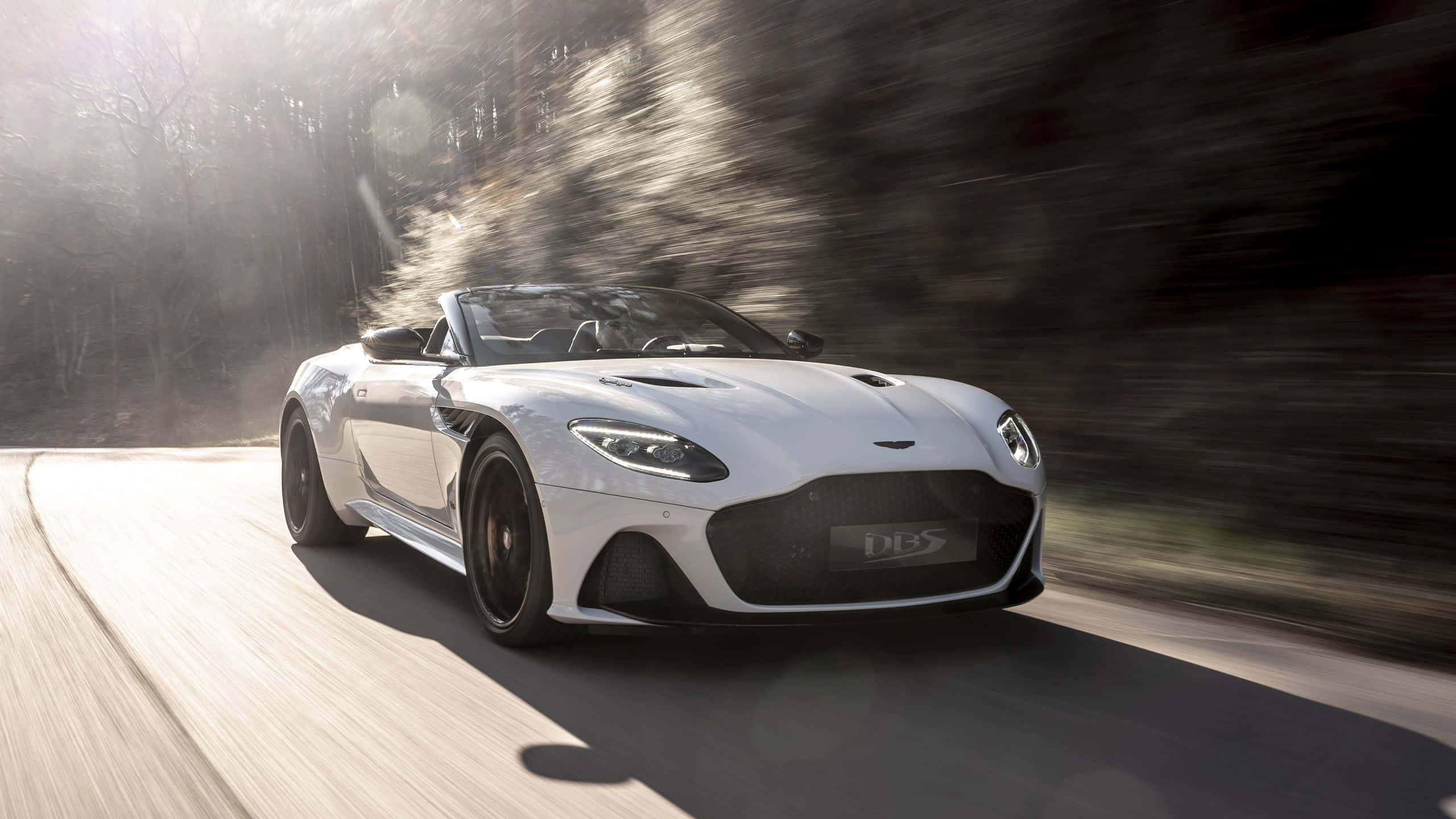Aston Martin Dbs Superleggera 桌布 壁紙 — 一架流線型白色Aston Martin DBS Superleggera敞篷跑車沿著蜿蜒的森林道路疾馳，動態模糊效果強調速度感。
