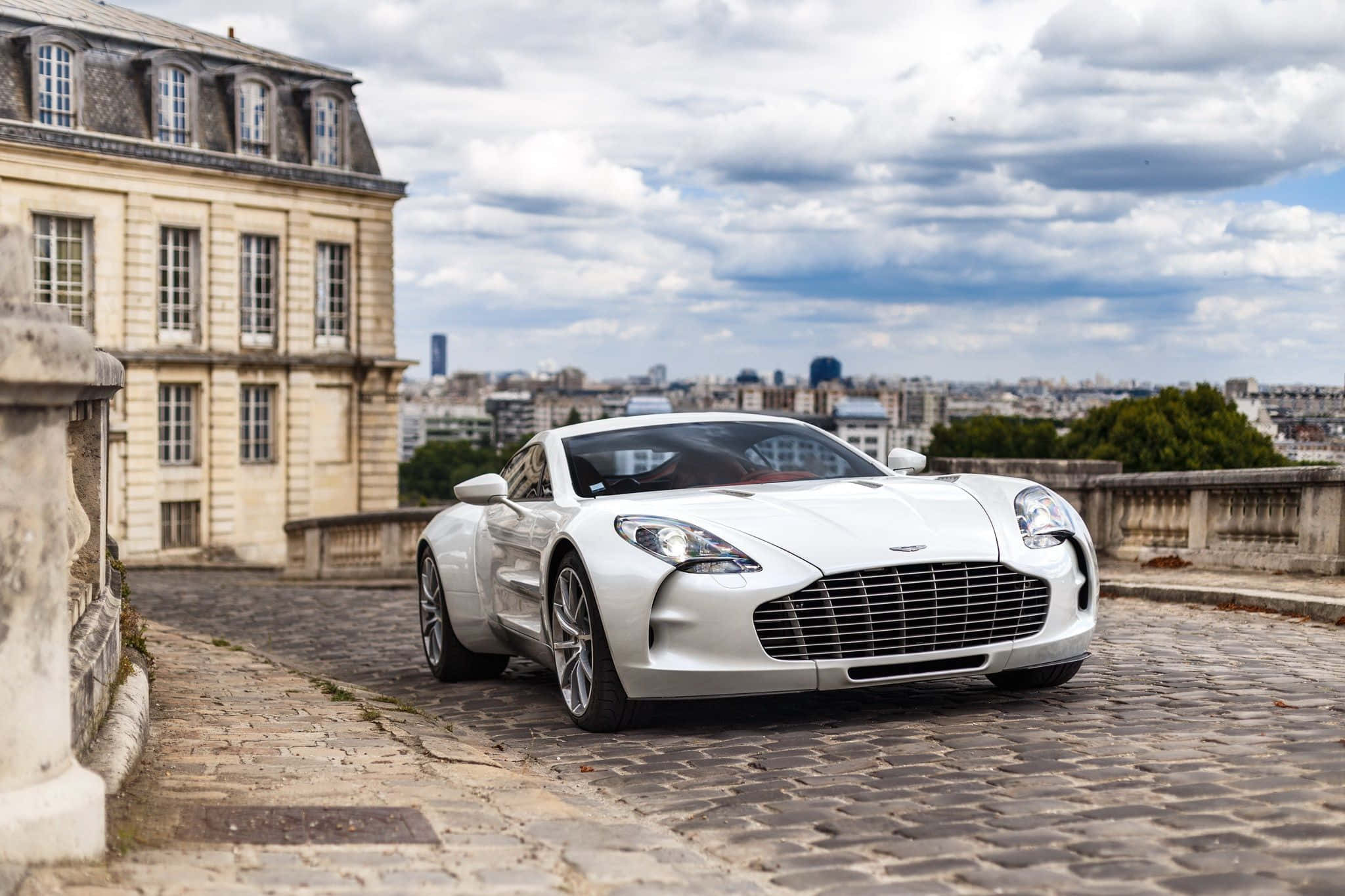 Aston Martin One-77 桌布 壁紙 — 一架流線型白色Aston Martin跑車停泊在鵝卵石屋頂上，俯瞰雲層下的巴黎城市景觀。