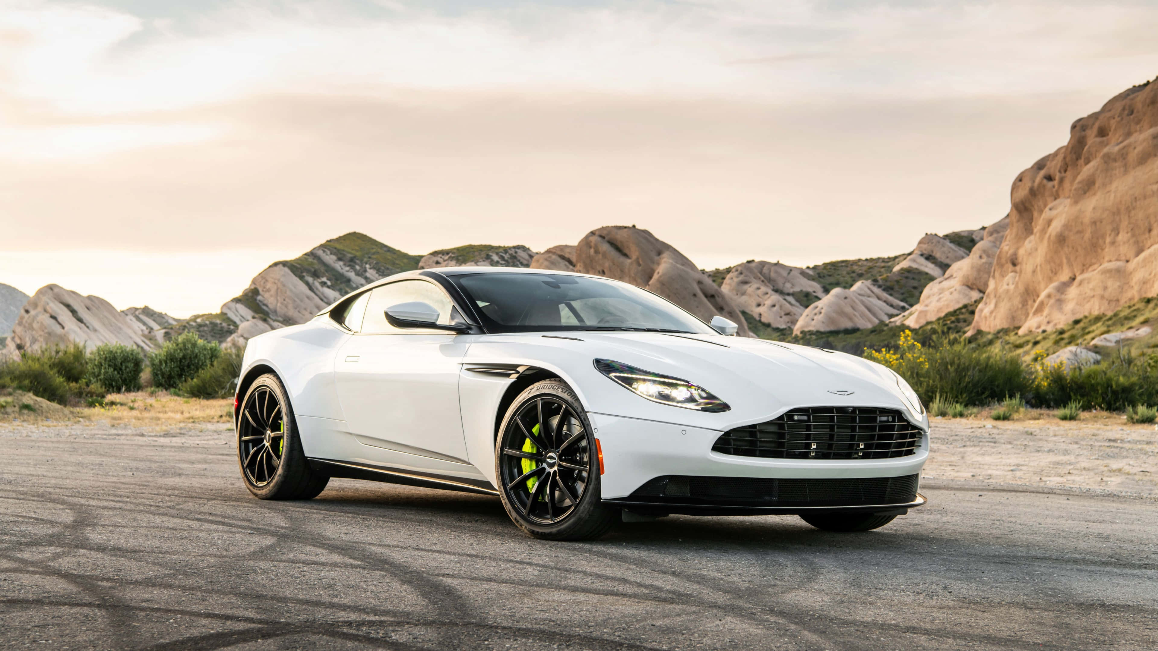 Aston Martin Db11 桌布 壁紙 — 一架流線型白色Aston Martin跑車停在沙漠公路上，周圍有岩石山丘，天空柔和金黃。