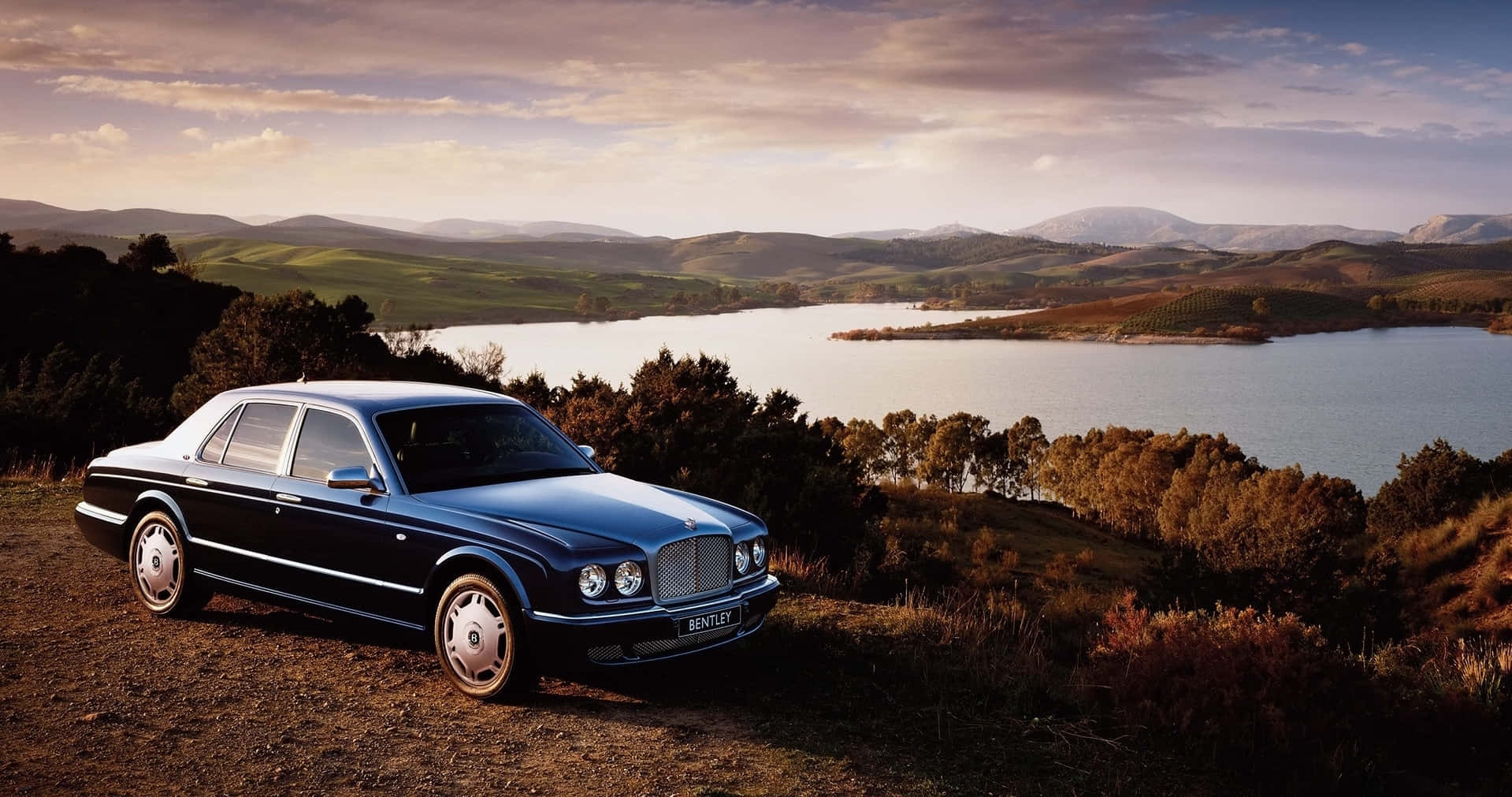 Bentley Arnage 桌布 壁紙 — 一輛流線型黑色賓利轎車停放在青草山坡上，俯瞰寧靜的湖泊和連綿起伏的丘陵，天空呈現柔和的金色日落。