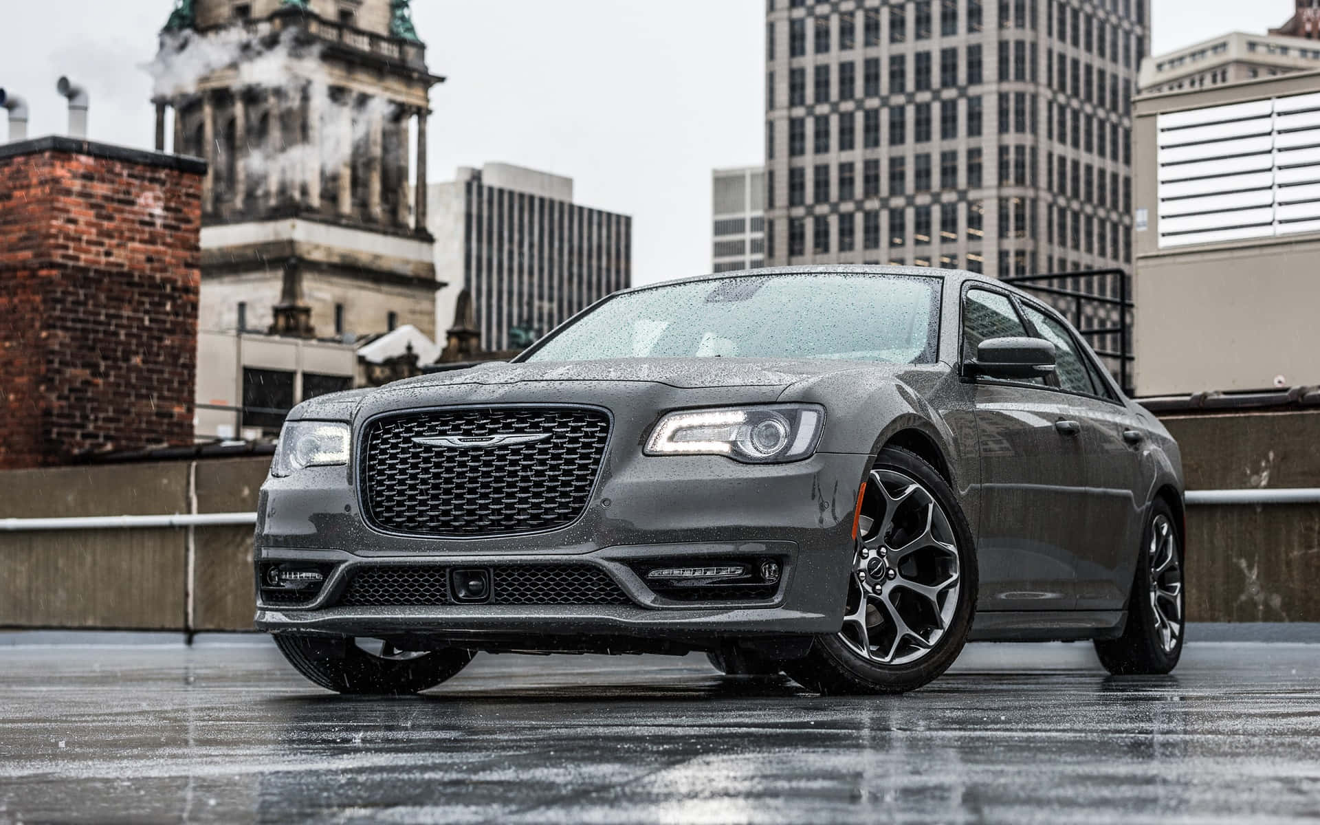Chrysler 300 桌布 壁紙 — 一架流線型灰色Chrysler 300轎車在潮濕的屋頂上閃閃發光，背景是城市摩天大樓和磚砌建築，天空多雲。