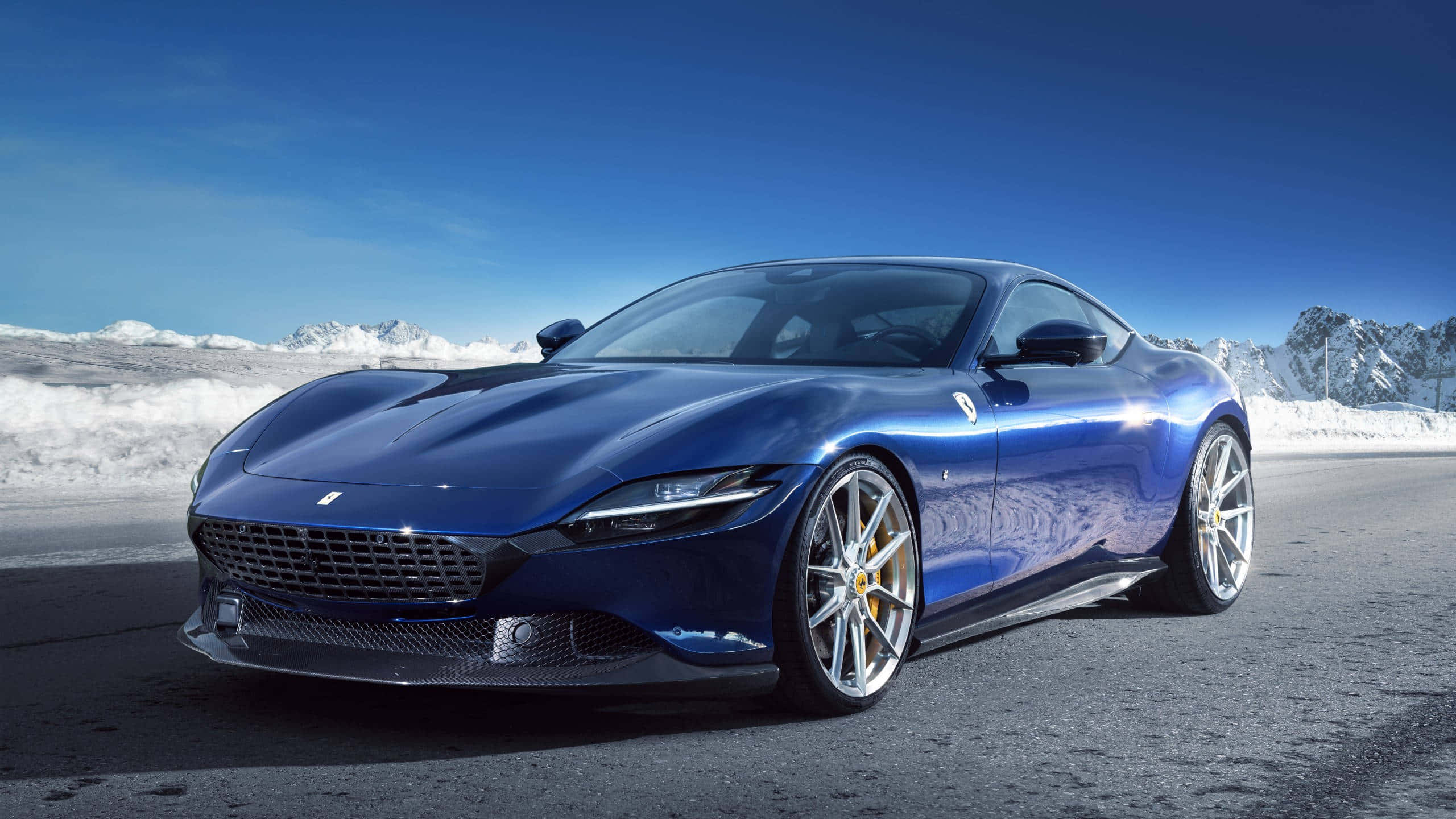 Ferrari Roma 桌布 壁紙 — 一輛流線型藍色法拉利跑車在晴朗藍天下閃耀於積雪的山路上。