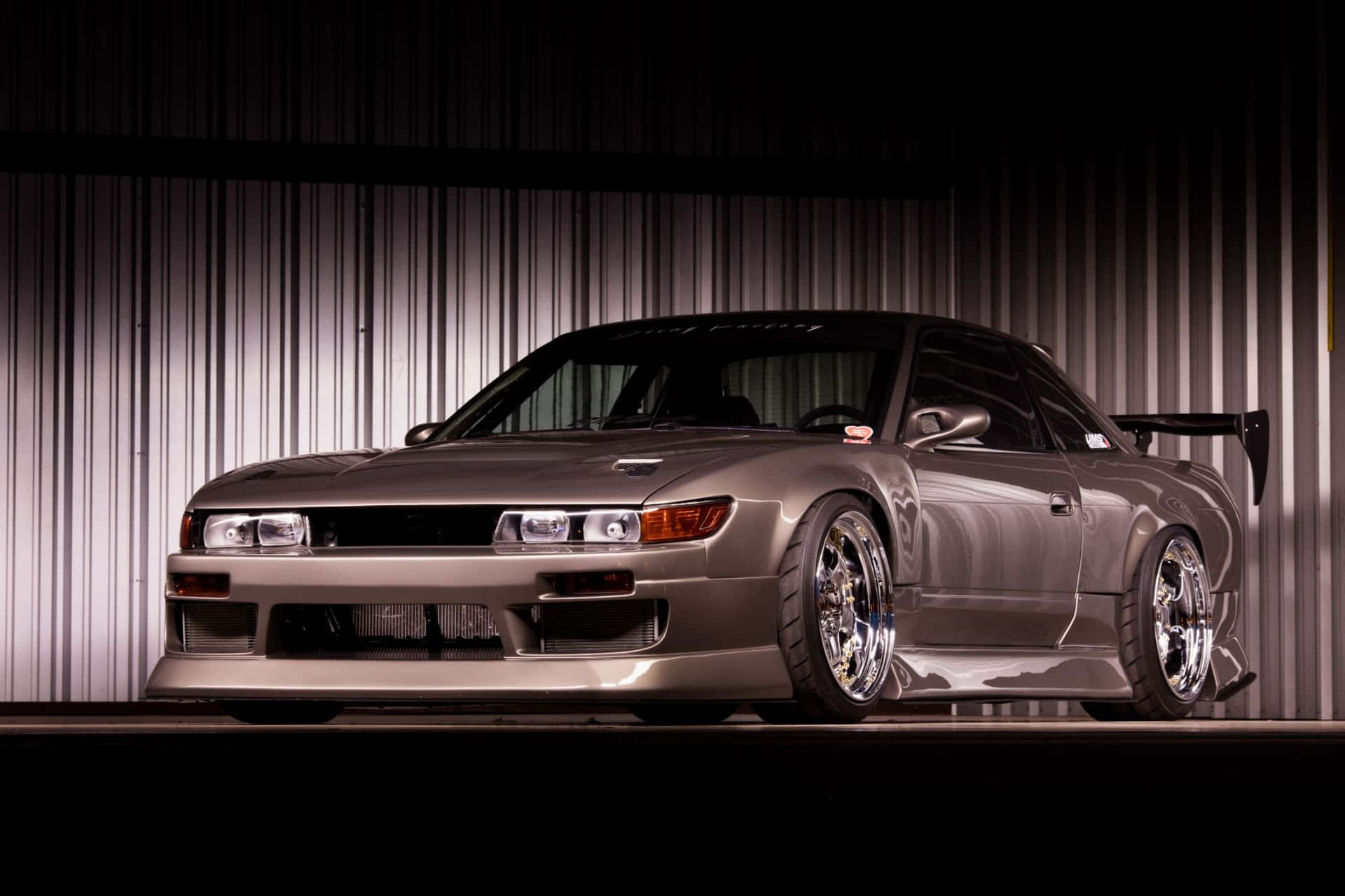  壁紙 — 一台流線型銀色日產Skyline GT-R配備定制寬體套件和鍍鉻輪圈，在波紋金屬牆前戲劇性地照亮。