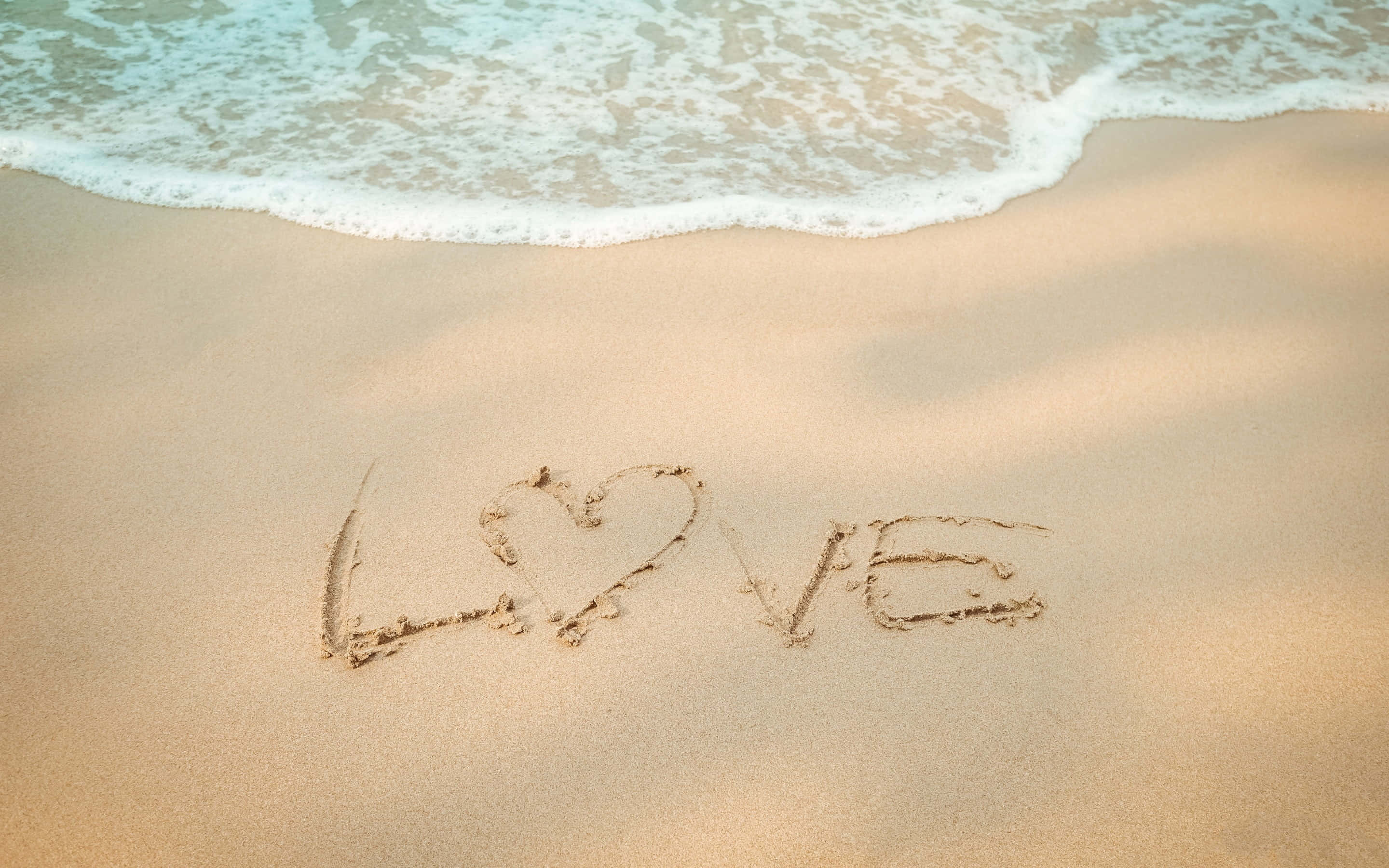 Breeze 桌布 壁紙 — 「LOVE」一詞中的O被心形取代，寫在濕沙上，溫柔的海浪緩緩湧入。
