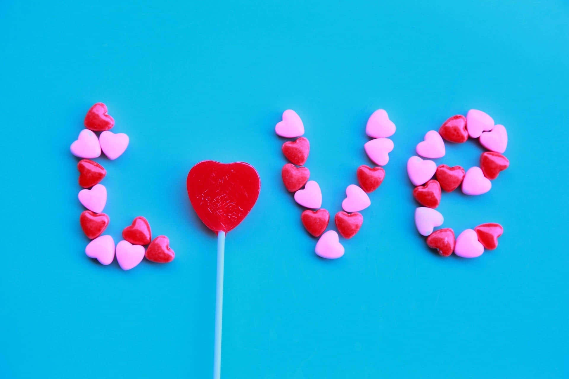 愛心 桌布 壁紙 — 「love」這個字用色彩繽紛的心形糖果和紅色心形棒棒糖在充滿活力的藍色背景上拼出來。