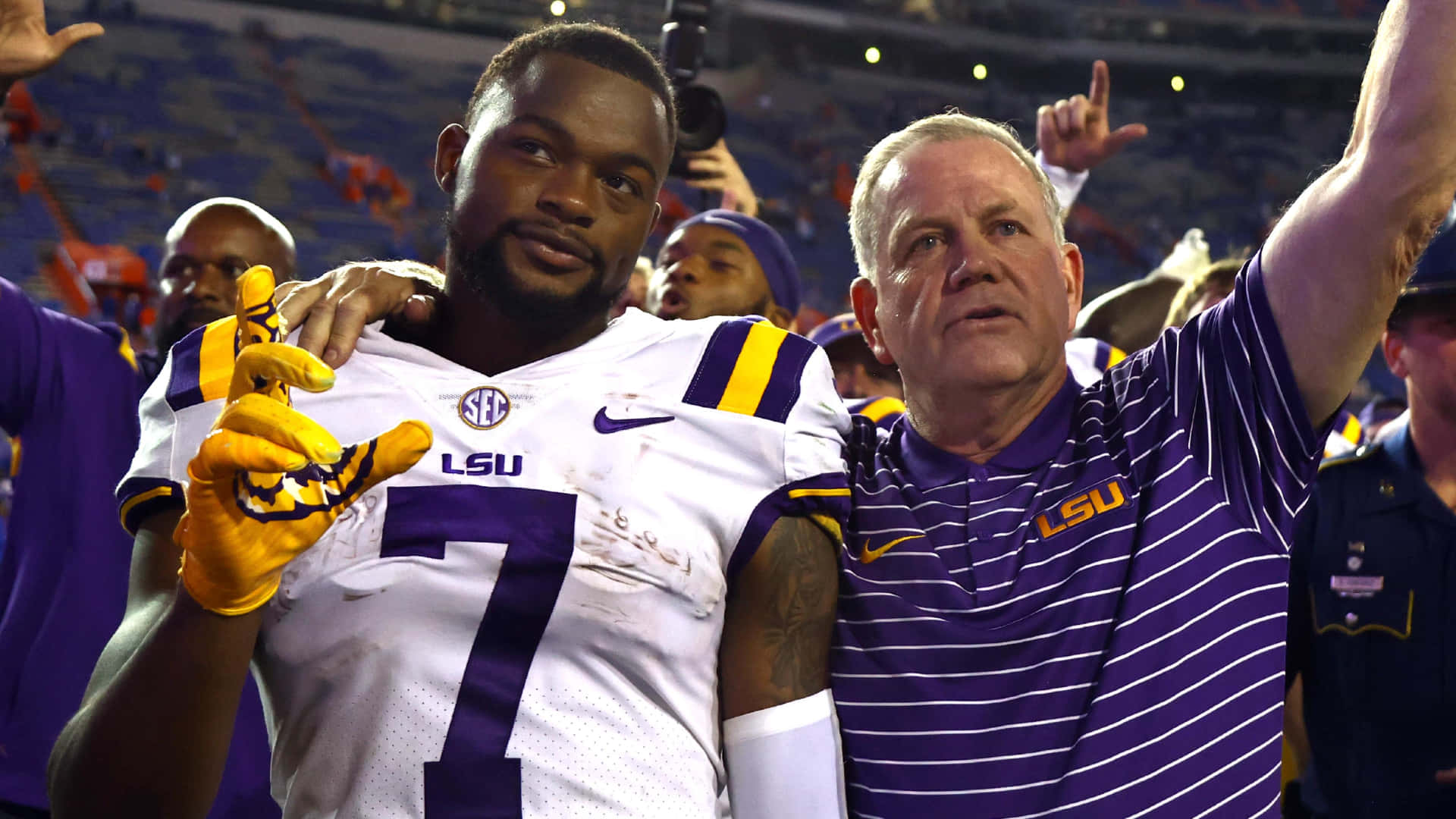  壁紙 — LSU 足球運動員 7 號身穿白色球衣配紫色點綴和黃色手套，在明亮燈光下的體育場與教練慶祝。