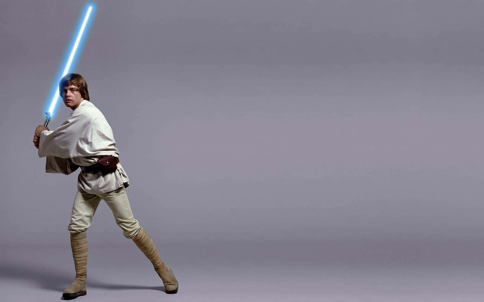 luke skywalker 桌布 壁紙 — Luke Skywalker 以戰鬥姿態站立，手持發光的藍色光劍，背景為純灰色攝影棚。