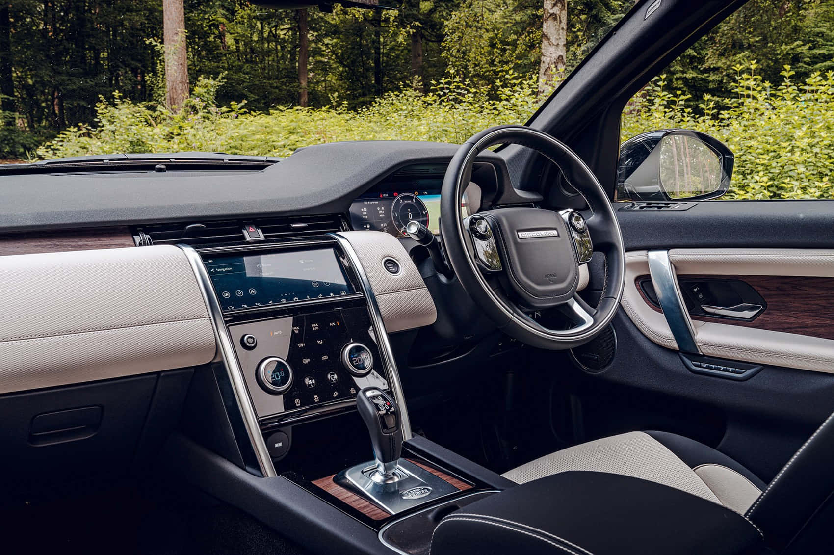 Land Rover Discovery 桌布 壁紙 — 豪華車內裝，搭配米色皮革和木質飾板，從駕駛座視角，車外有森林。