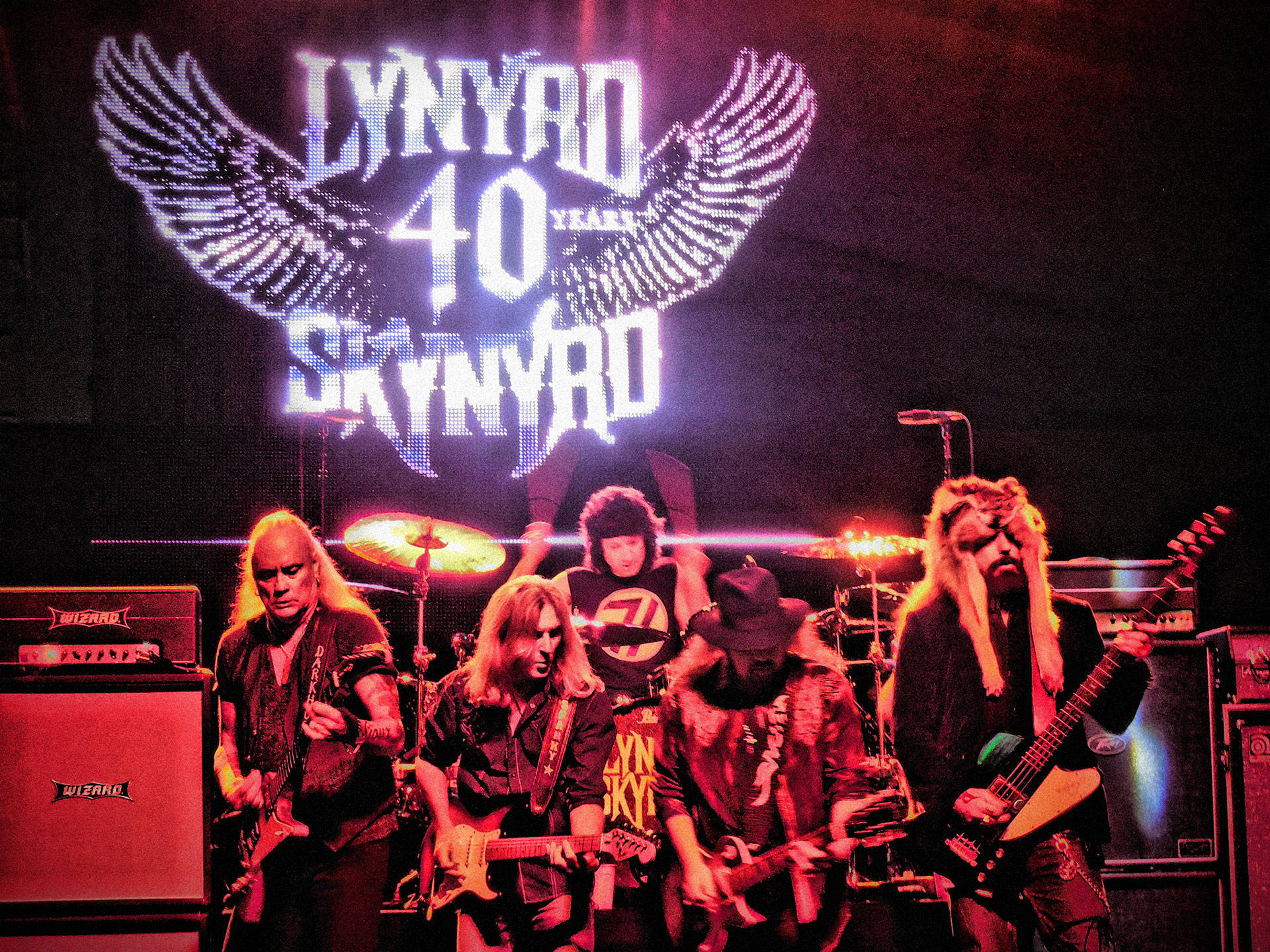 音樂 桌布 壁紙 — Lynyrd Skynyrd 樂團在舞台上現場表演，背景是發光的 '40 Years' 翅膀標誌，紅色舞台燈光和吉他。