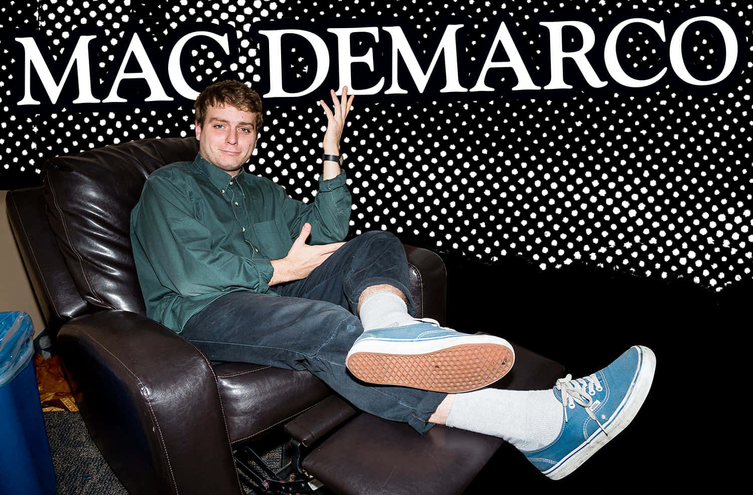  壁紙 — Mac DeMarco 在黑色牆壁搭配白色波點的背景下，隨意地躺在皮製躺椅上，他的名字以粗體白色字呈現。