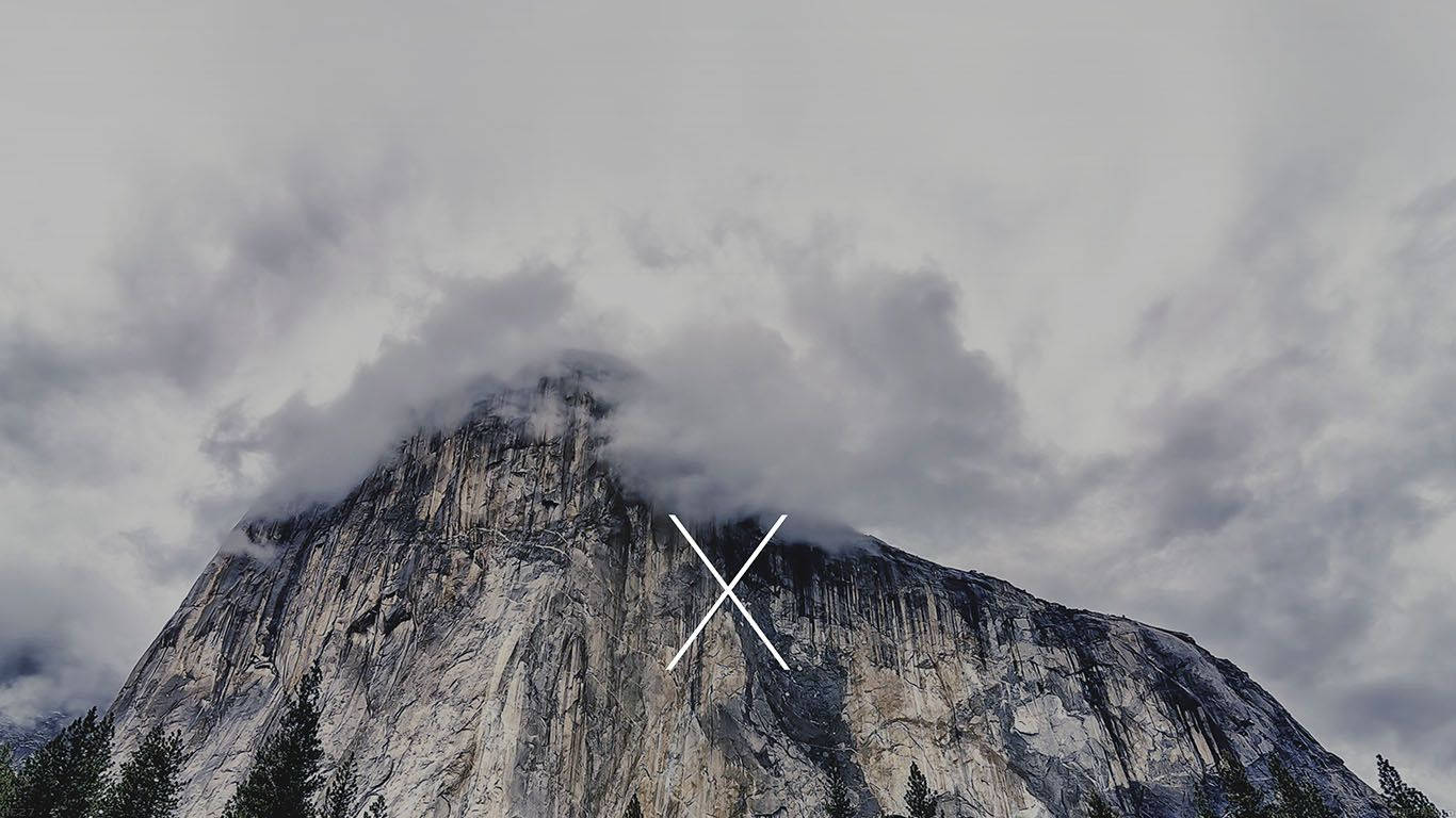 從您的新MacBook欣賞優勝美地山脈的壯麗景色 壁紙 — 一座高聳的花崗岩山峰，部分被戲劇性的灰色雲霧籠罩，山腳有常綠樹木。