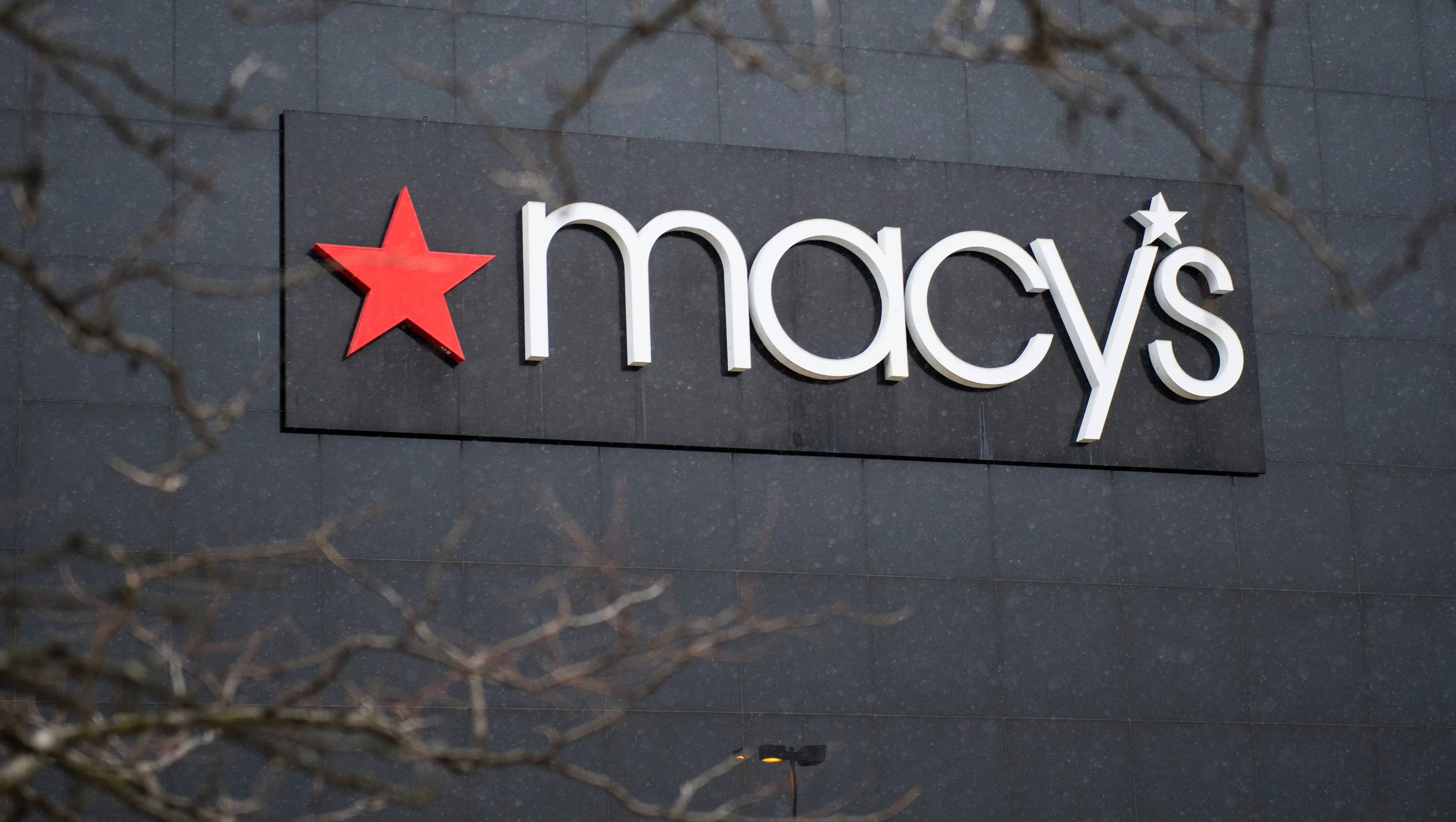 macys 桌布 壁紙 — Macy's 商店標誌的特寫，紅色星星和白色字體安裝在深灰色瓷磚牆上，由光禿的樹枝框住。