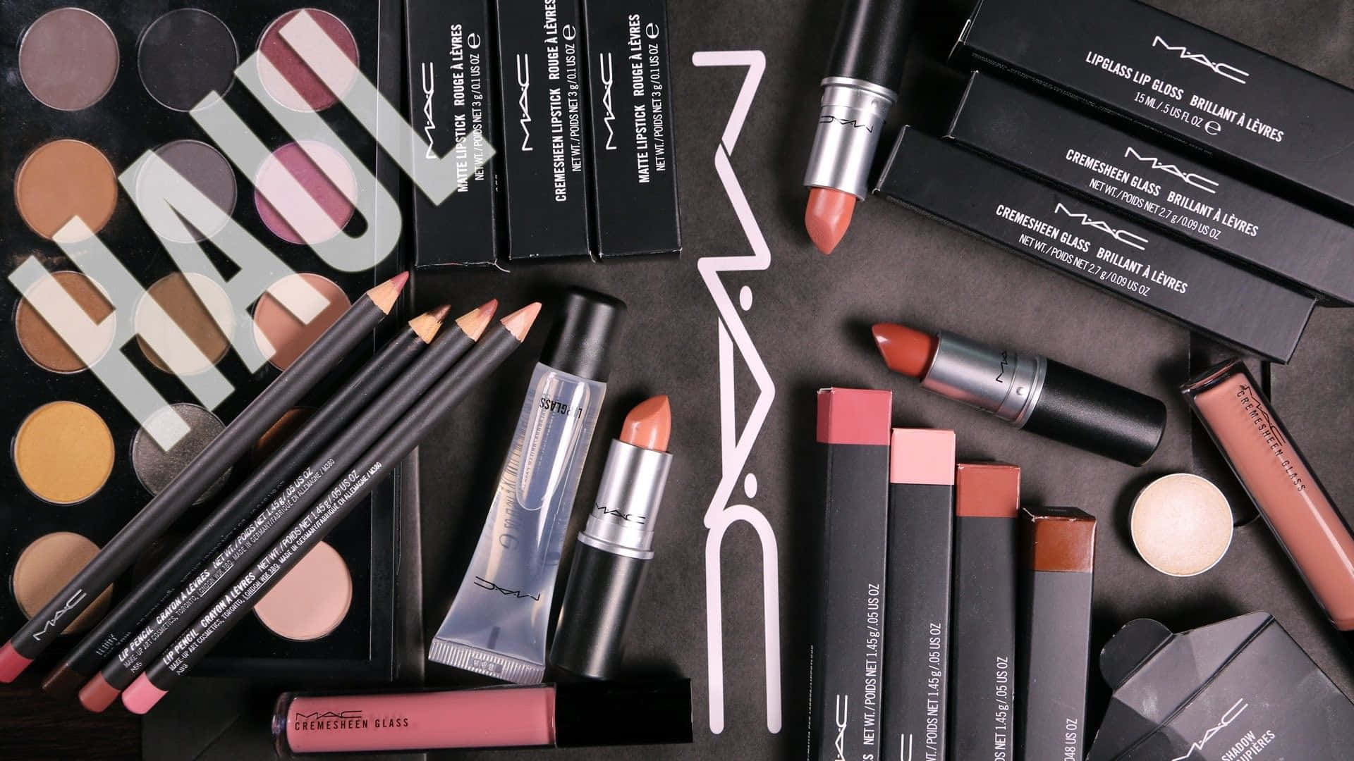 Mac Cosmetics 桌布 壁紙 — MAC化妝品（包括唇膏、眼線筆和眼影盤）在深色表面上平鋪擺放，帶有大型白色「HAUL」浮水印。
