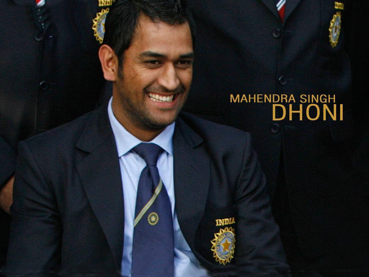 Ms Dhoni 桌布 壁紙 — Mahendra Singh Dhoni 身穿深色西裝，帶有印度板球隊徽章，背景是模糊的球隊背景，容光煥發。