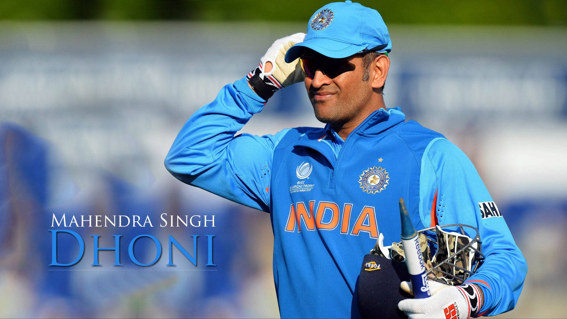MS Dhoni 桌布 壁紙 — Mahendra Singh Dhoni 身穿藍色印度板球裝備，在陽光明媚的場地上手持球棒和頭盔，輕觸帽沿。
