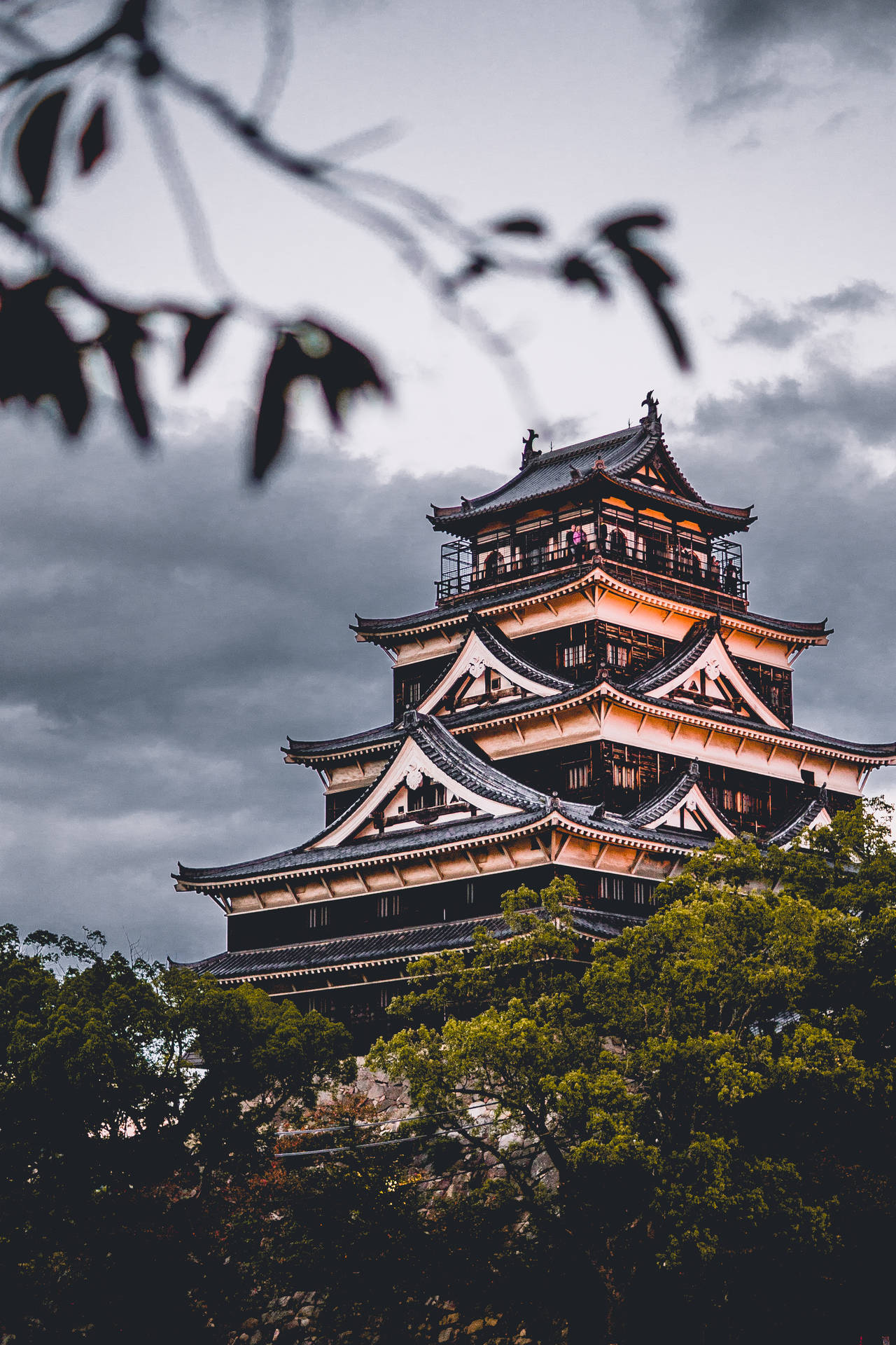 一座歷史悠久的日本城堡在多雲的天空下溫暖地發光，前景有樹木。 — 日式 壁紙