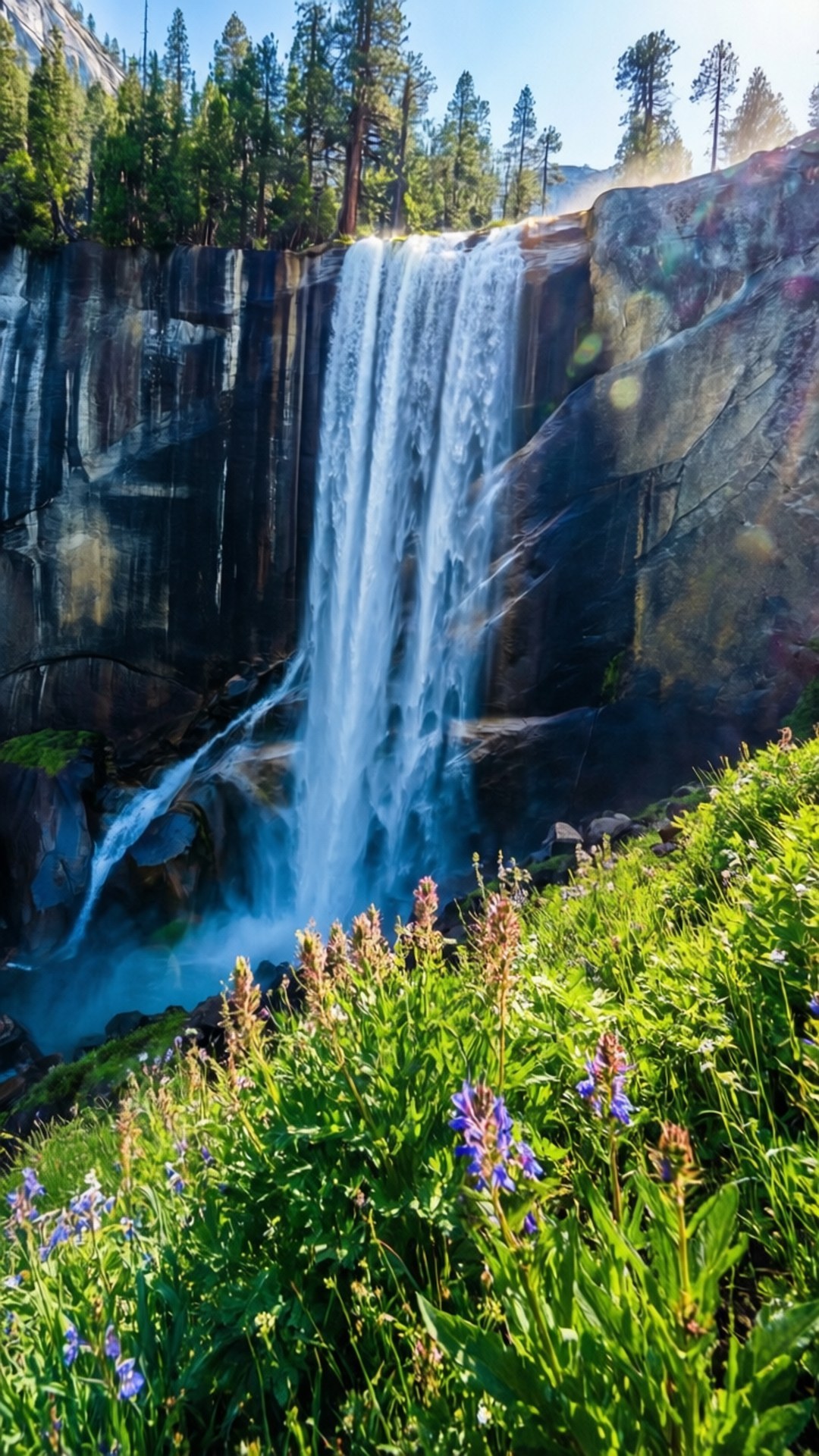 Spring Yosemite Falls 壁紙 — 一道雄偉的瀑布從崎嶇的懸崖峭壁傾瀉而下，周圍環繞著高聳的常青樹和前景中充滿活力的紫色野花。