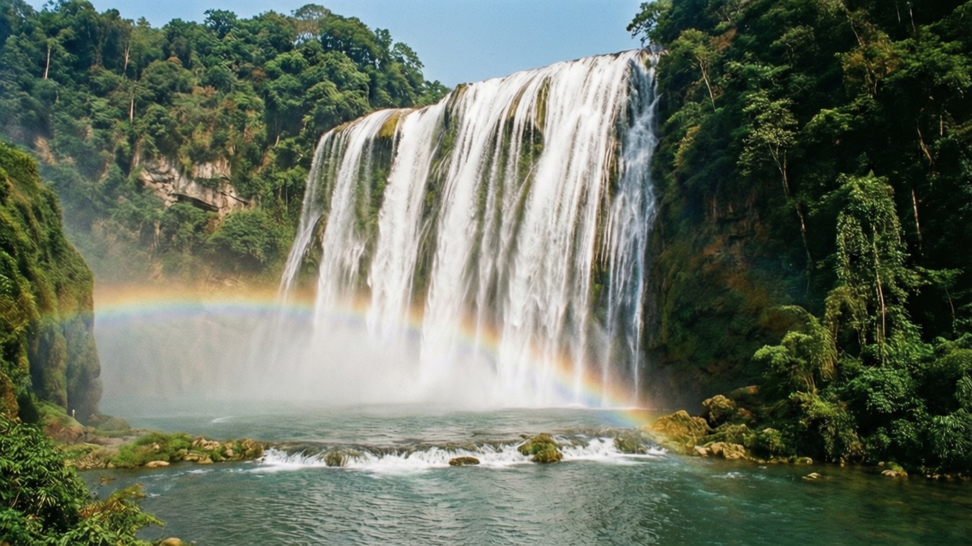 Waterfall Rainbow Peace 壁紙 — 一道雄偉的寬瀑布穿過茂密的綠色叢林，在下方霧氣瀰漫的水潭上形成一道鮮豔的彩虹。
