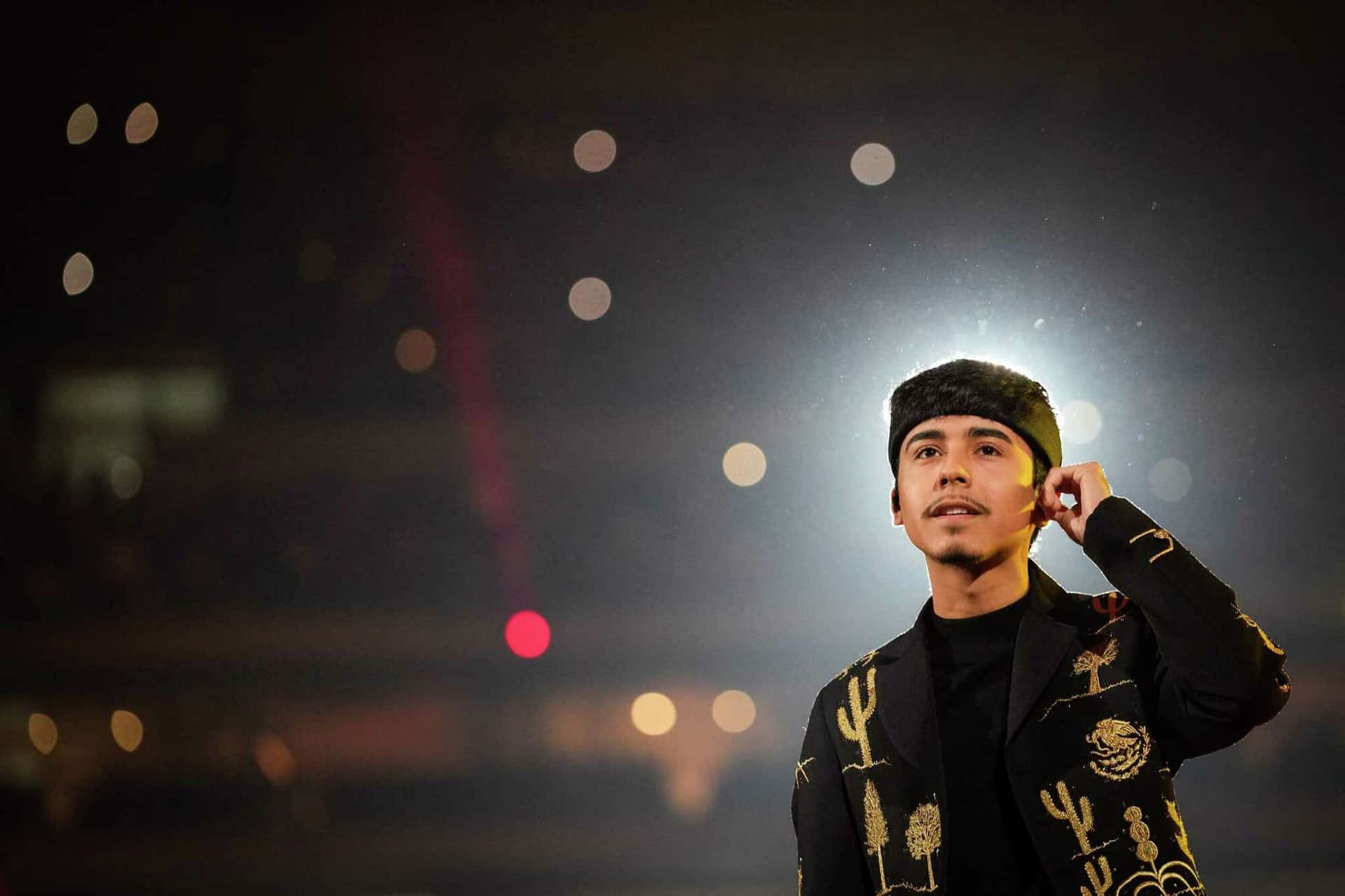  壁紙 — 一名身穿黑色仙人掌圖案外套和毛帽的男子，站在模糊的夜景城市背景前，背景有發光的散景燈光。