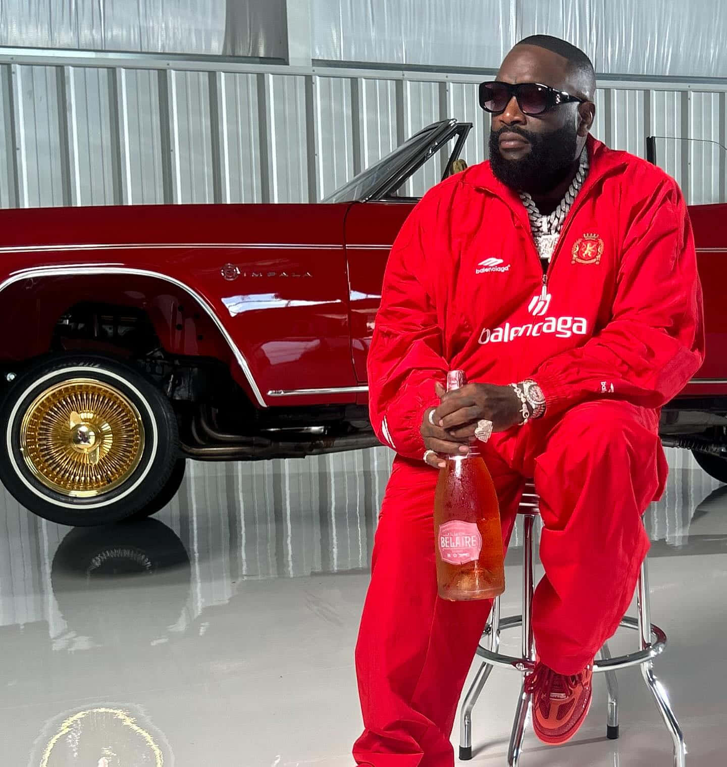 壁紙 — 一名身穿紅色Balenciaga運動服的男子坐在凳子上，在車庫裡拿著一瓶香檳，旁邊是一輛閃亮的紅色經典汽車，配有金色輪圈。