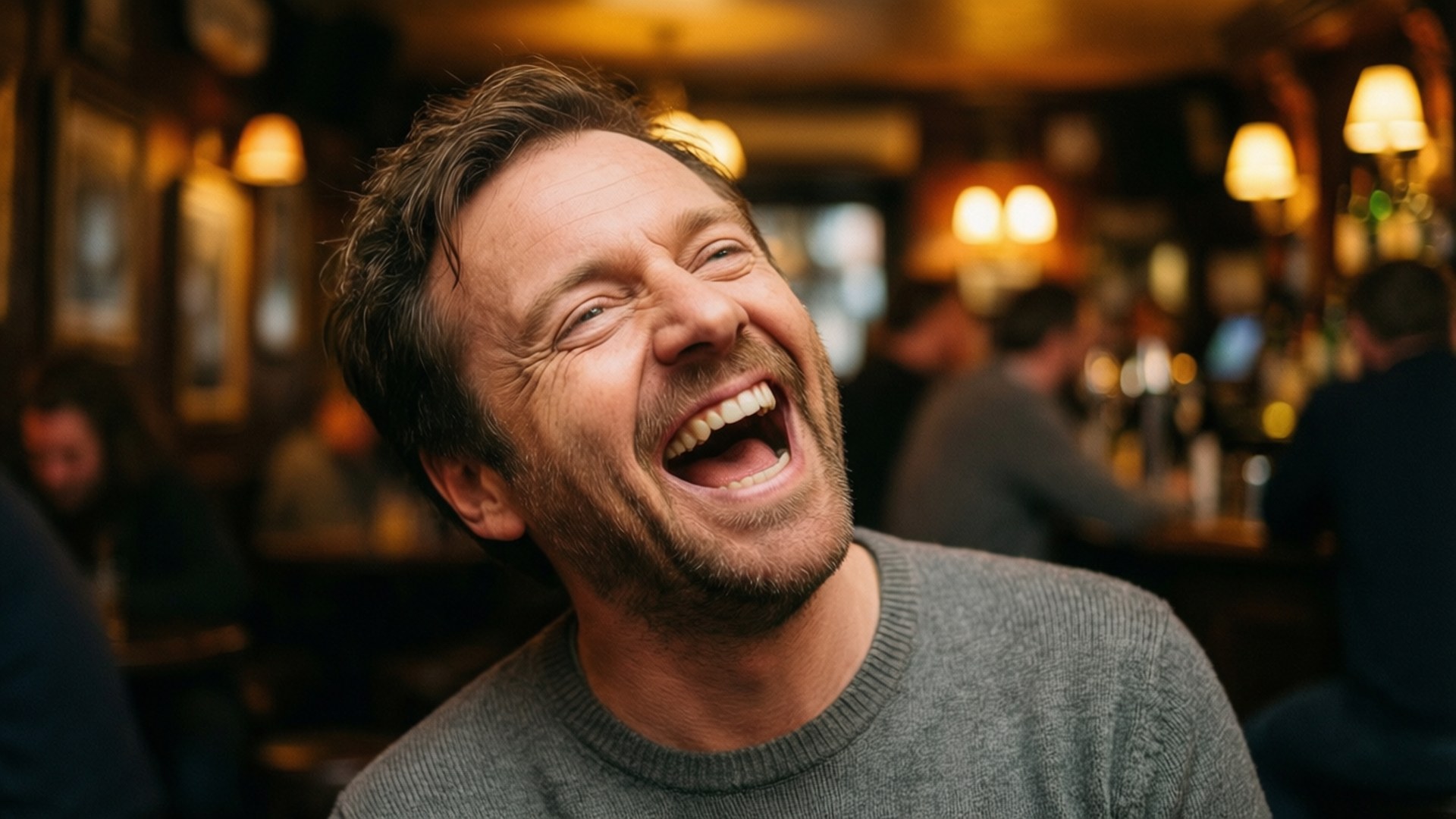 ricky gervais 桌布 壁紙 — 一個男人在溫暖燈光的酒吧裡，木牆和吊燈下開懷大笑。