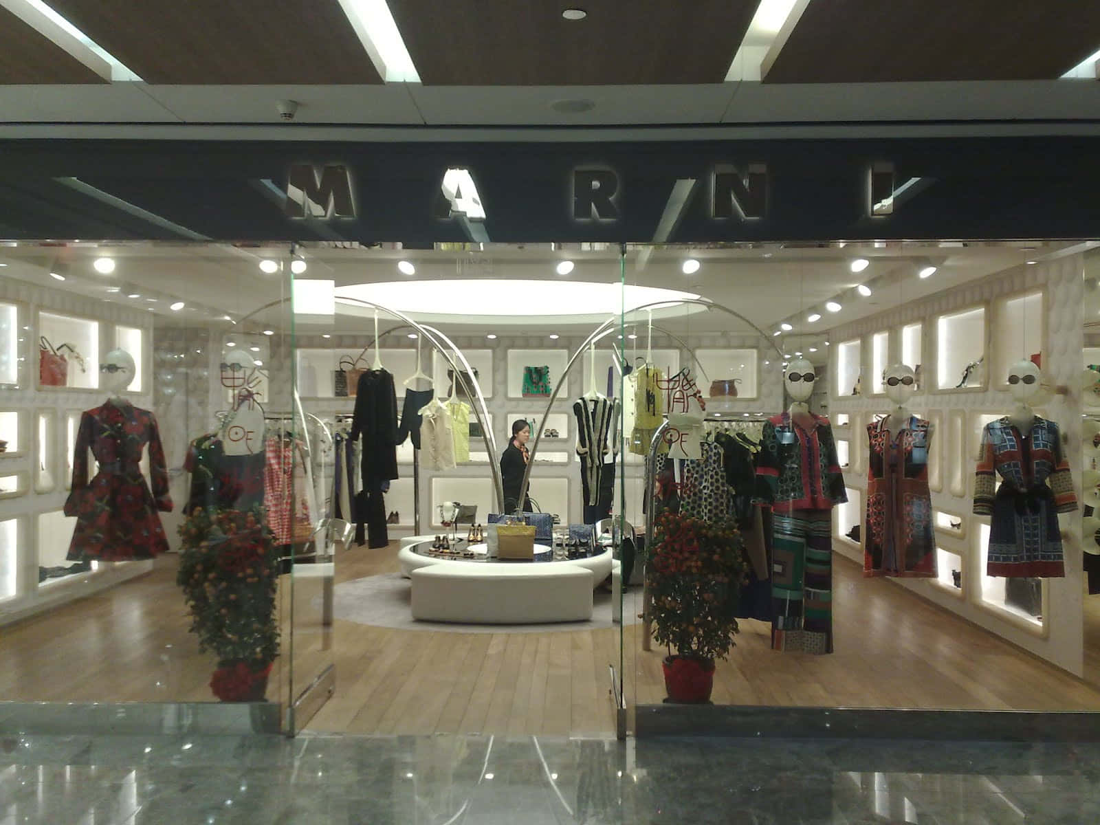 Store 桌布 壁紙 — Marini 時裝精品店在明亮的燈光下展示穿著彩色圖案連衣裙的人體模型，入口兩側擺放盆栽植物。