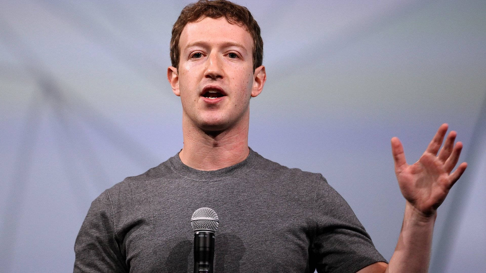 商業 桌布 壁紙 — Mark Zuckerberg 對著麥克風說話，做出張開手勢，背景是柔和的藍色。