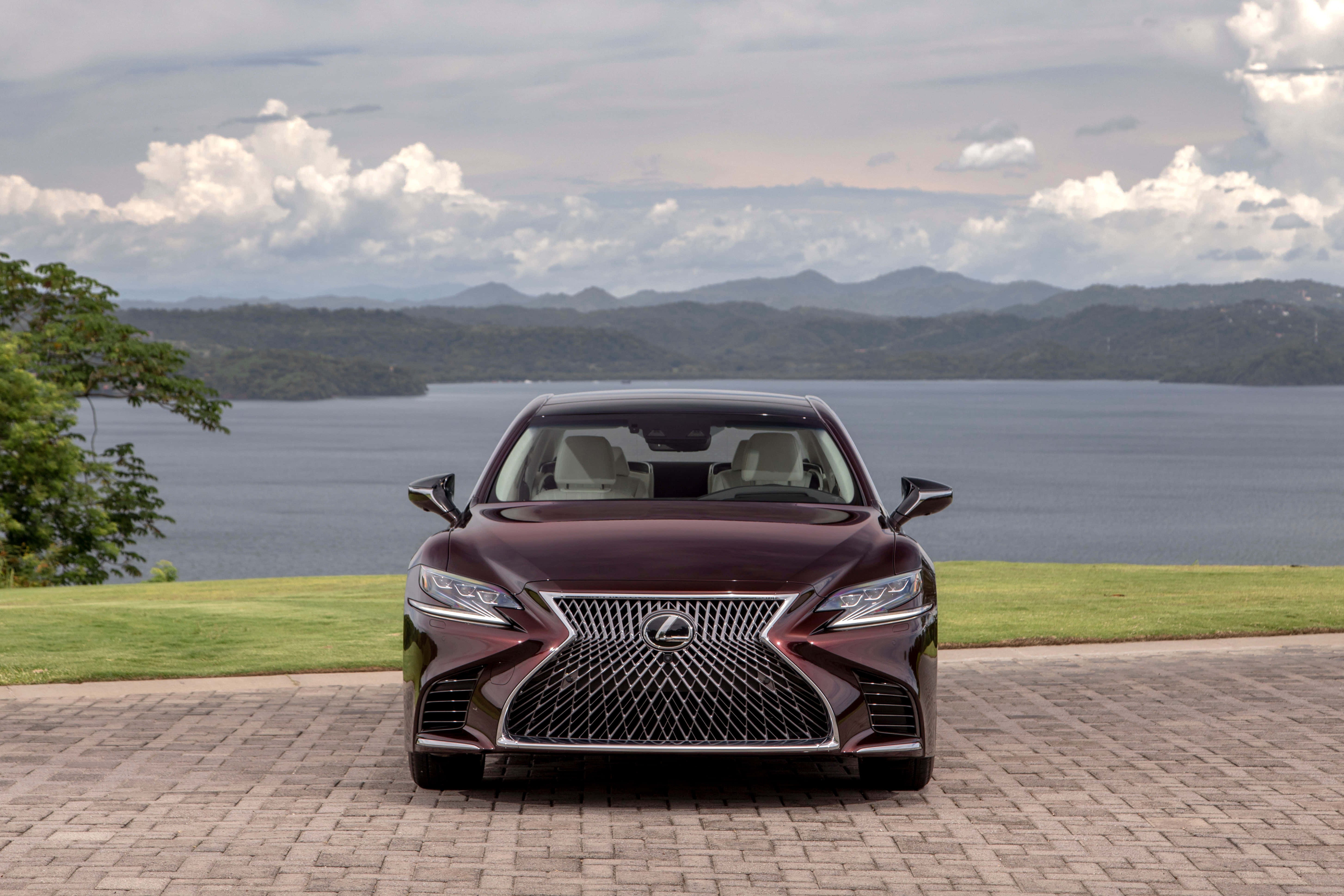 Lexus Ls 500 桌布 壁紙 — 一輛栗色Lexus轎車在磚砌車道上向前行駛，前方是廣闊的湖泊和森林覆蓋的山脈，天空多雲。