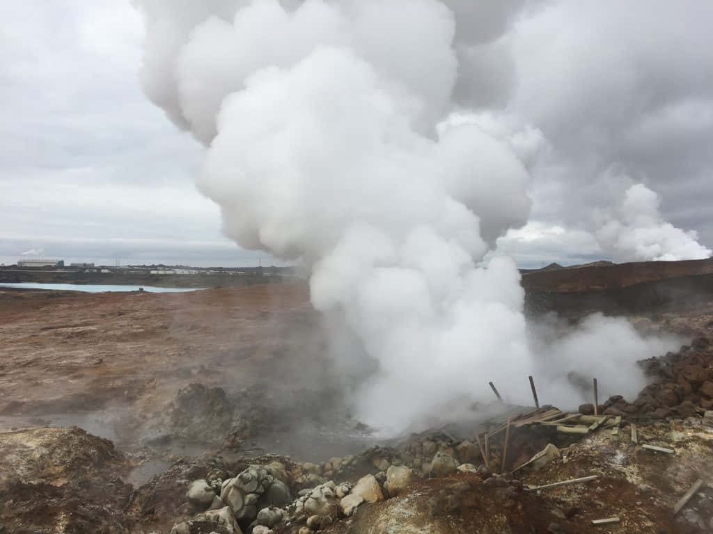  壁紙 — 在陰天之下，崎嶇的火山地貌中，一個地熱通風口噴出巨大的白色蒸汽柱。