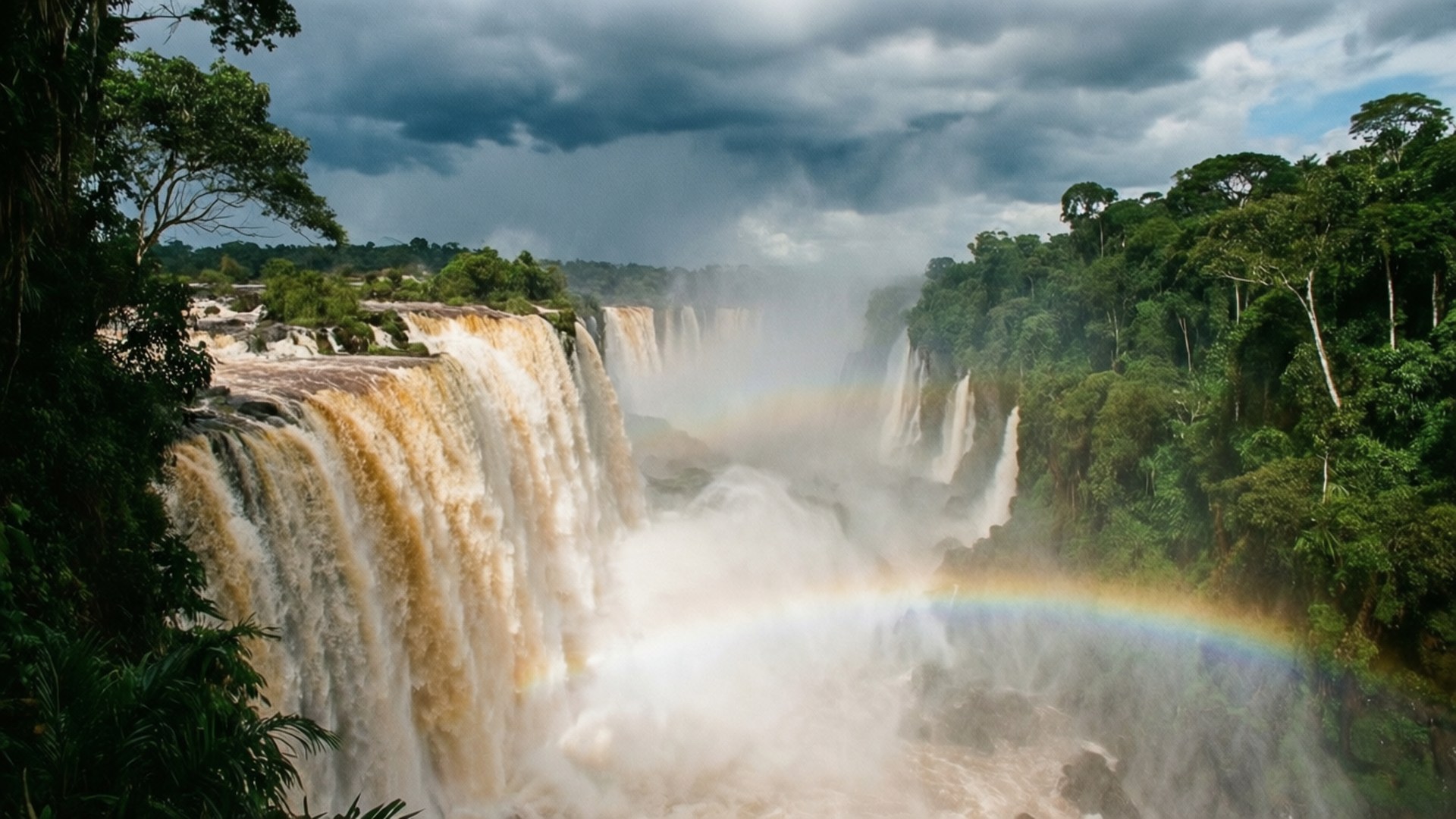 Iguazu Falls Power 壁紙 — 巨大的瀑布穿過茂密的綠色叢林傾瀉而下，在暴風雨的天空下，一道鮮豔的彩虹拱起在霧氣騰騰的水霧之上。
