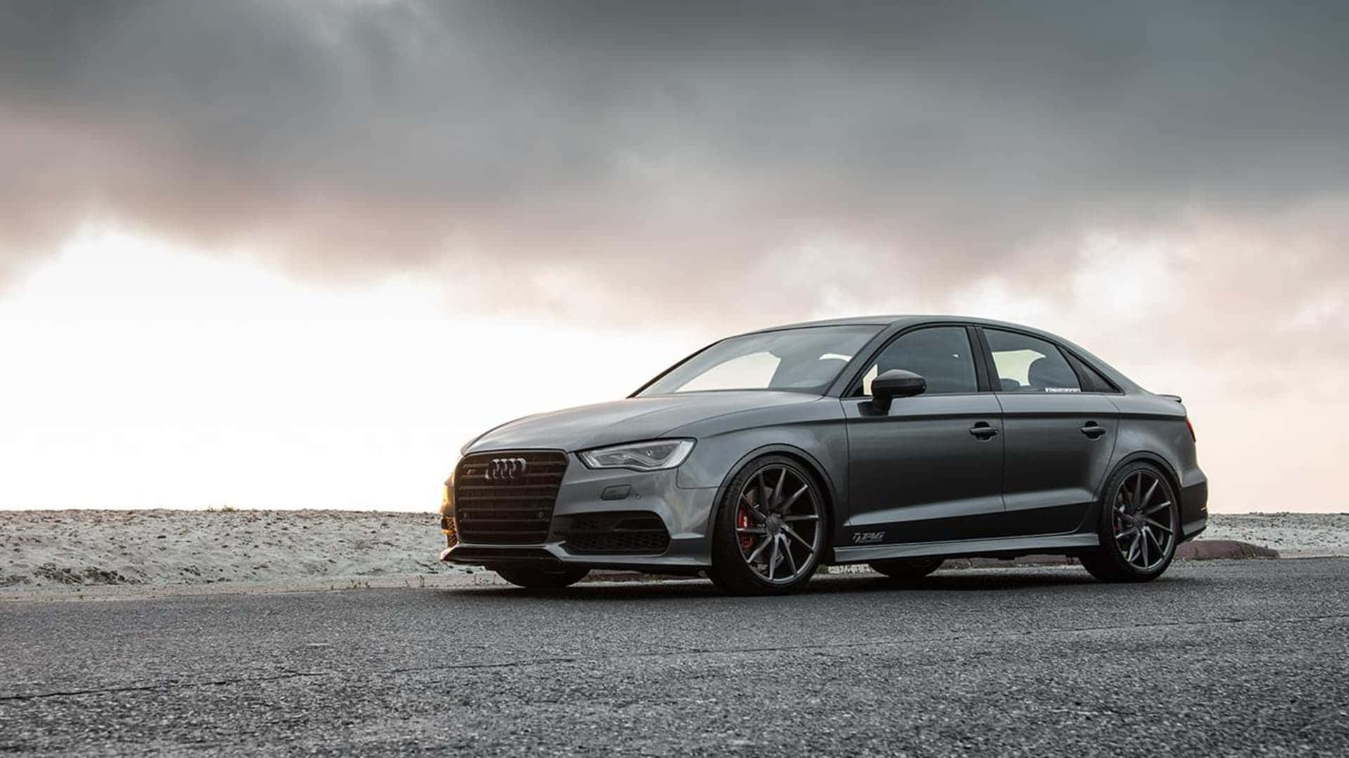 Audi S3 桌布 壁紙 — 一輛啞光灰色奧迪轎車搭配黑色輪圈，在黃昏時分戲劇性的多雲天空下，停在裂開的柏油路上。