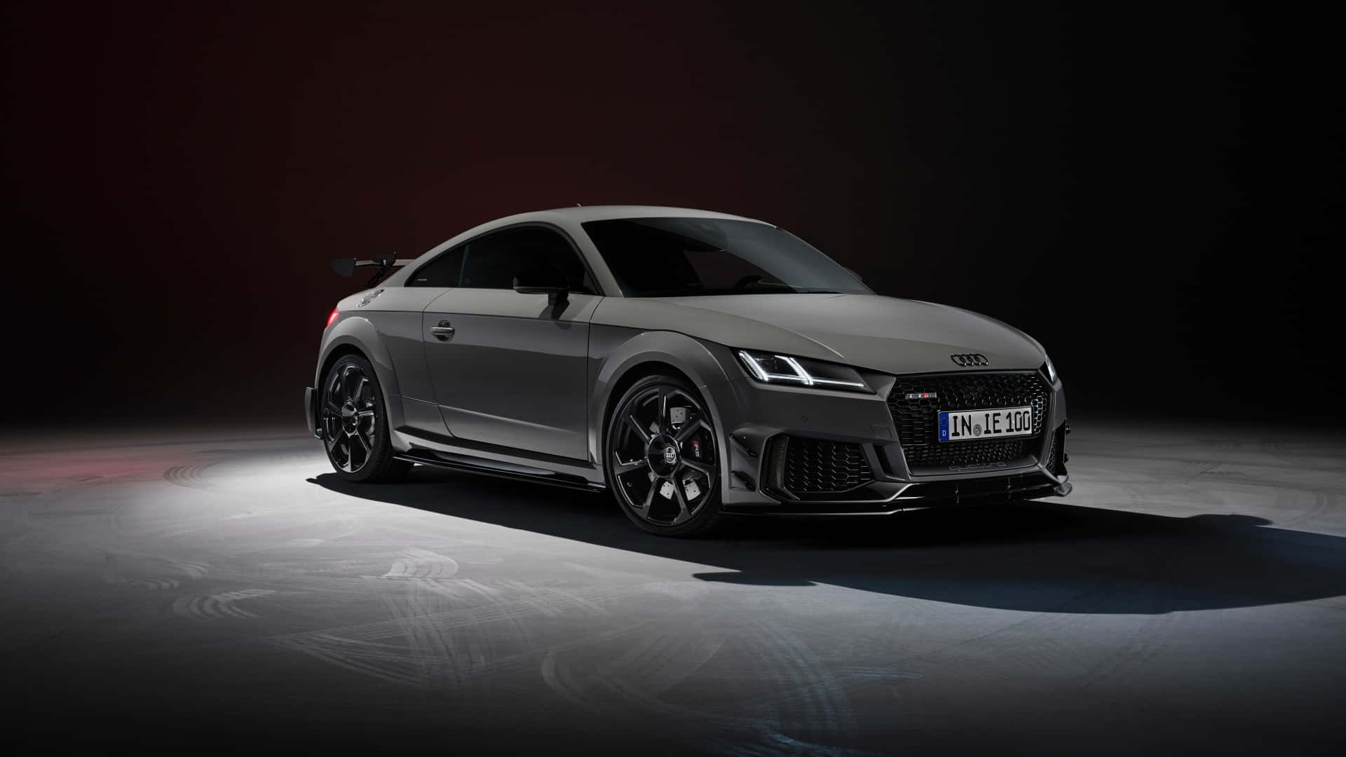 Audi Tt Rs 桌布 壁紙 — 一輛時尚的啞光灰色奧迪TT RS跑車在黑暗的攝影棚中，被紅色環境燈光和地板上的輪胎痕跡戲劇性地照亮。