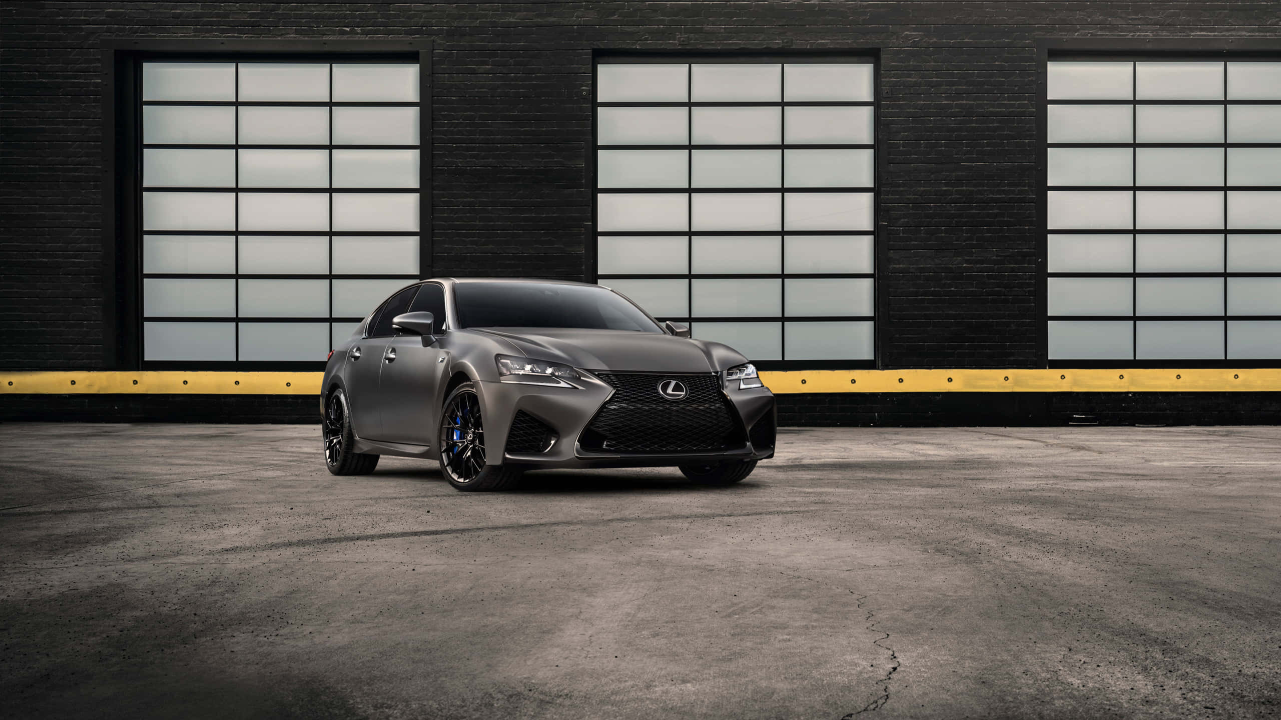 Lexus Gs 桌布 壁紙 — 一輛線條流暢的啞光灰色Lexus轎車搭配黑色輪圈，停在一間有大型窗戶的深色磚造倉庫前。