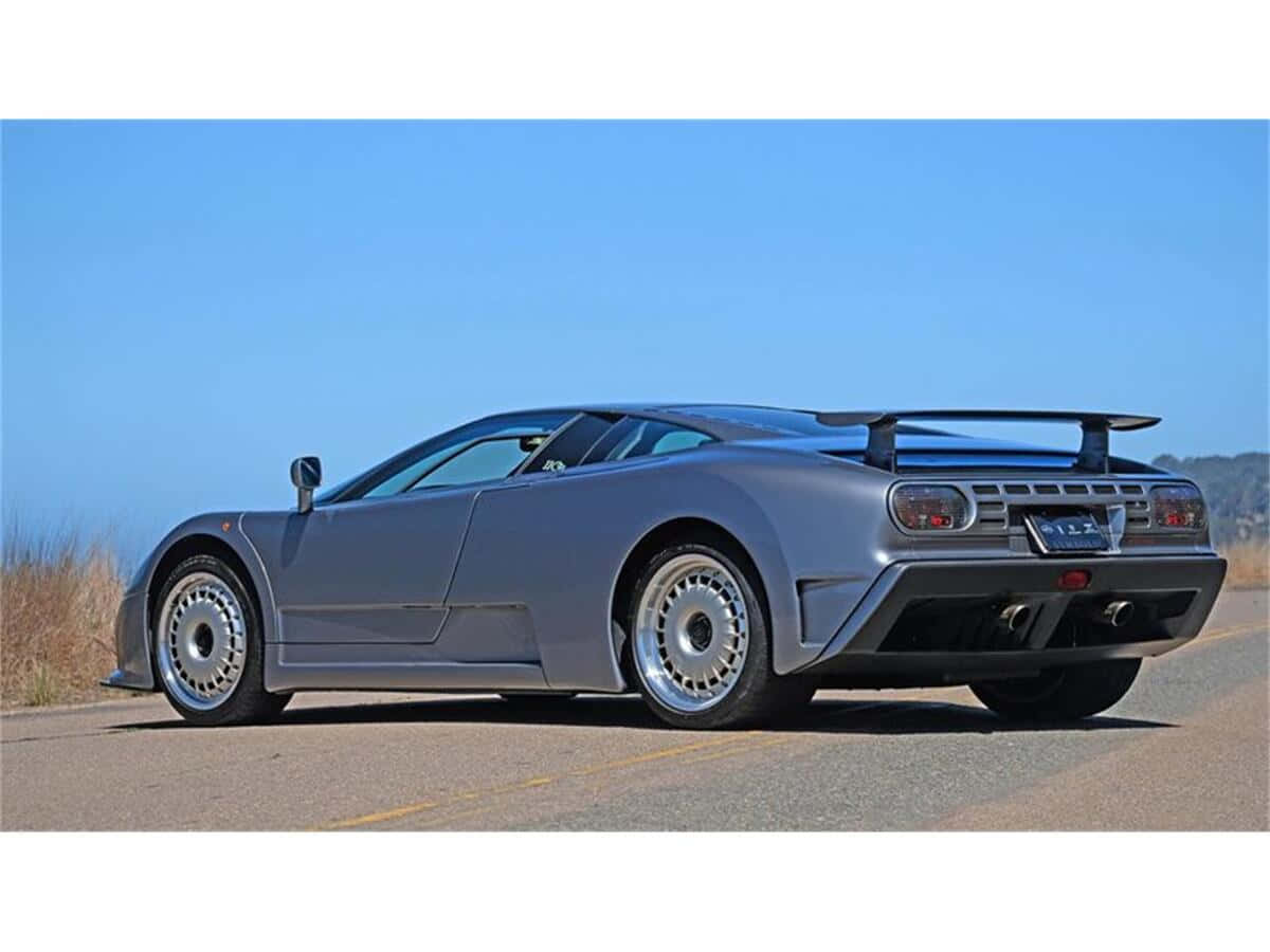 Bugatti Eb110 桌布 壁紙 — 一輛流線型啞光灰色SSC Ultimate Aero跑車停泊在陽光明媚的瀝青路上，周圍有乾草和山丘，天空清澈湛藍。