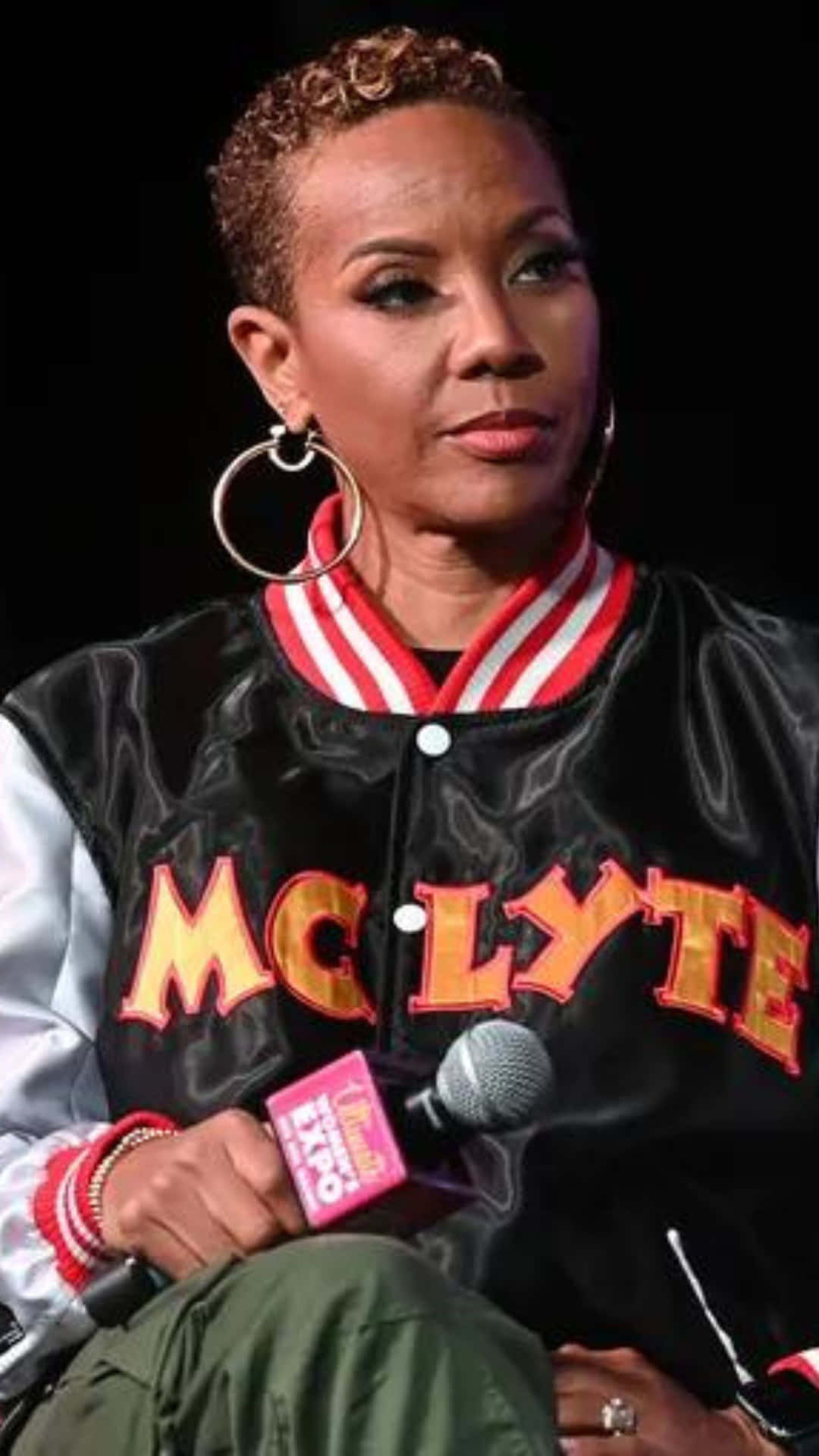 壁紙 — MC Lyte 自信地坐在活動中，身穿帶有紅白飾邊的黑色緞面外套，手持麥克風。