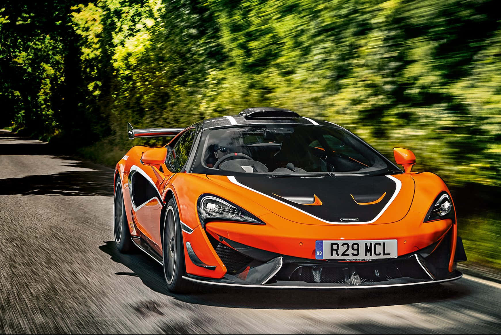 Mclaren 620r 桌布 壁紙 — 一輛充滿活力的橙色McLaren跑車在陽光明媚的森林道路上疾馳，茂密的綠樹呈現動態模糊效果。