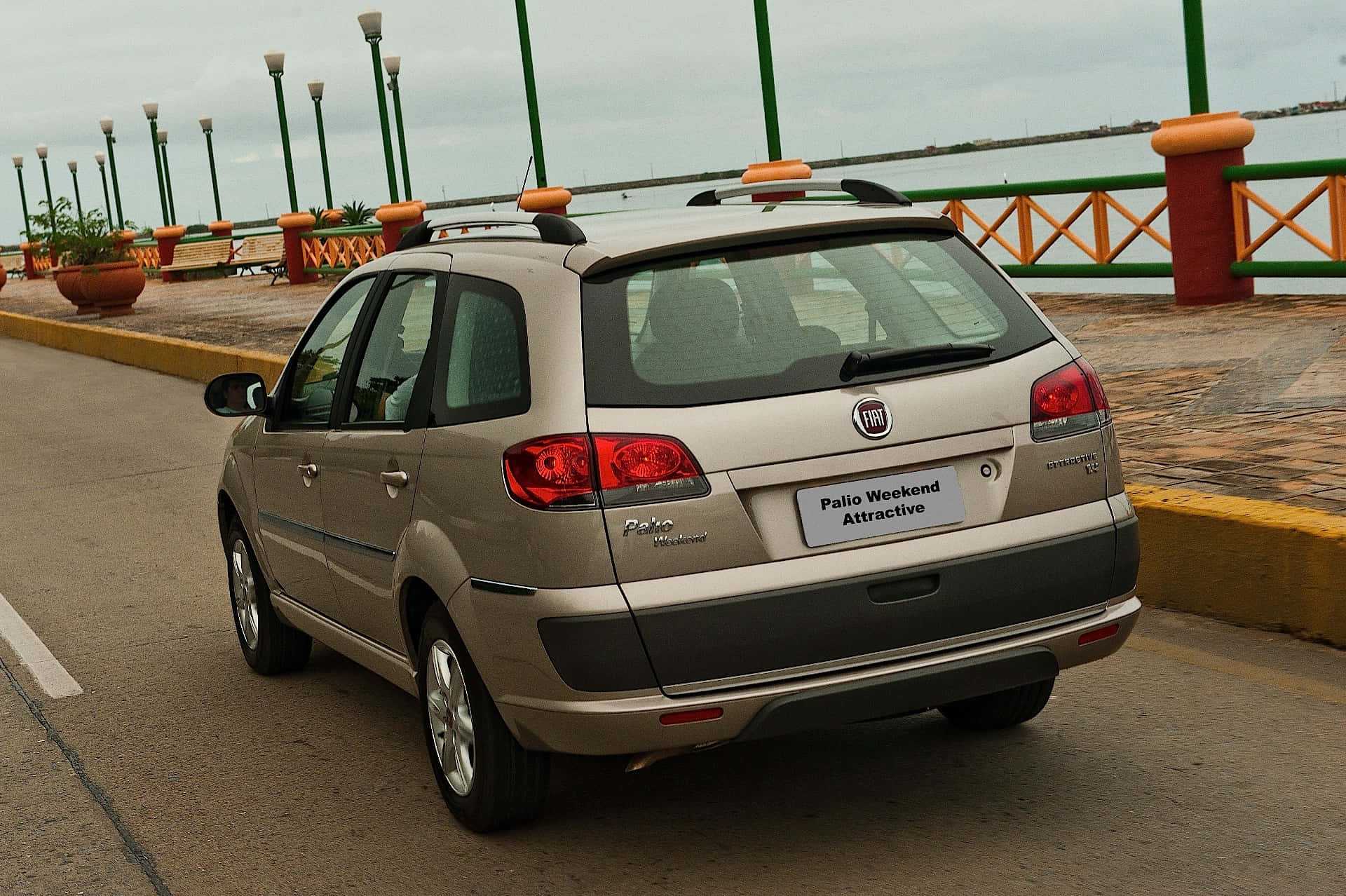 Fiat Palio 桌布 壁紙 — 一輛米色Fiat Palio Weekend Attractive旅行車停放在海濱長廊上，周圍有色彩繽紛的欄杆和盆栽植物，天空多雲。
