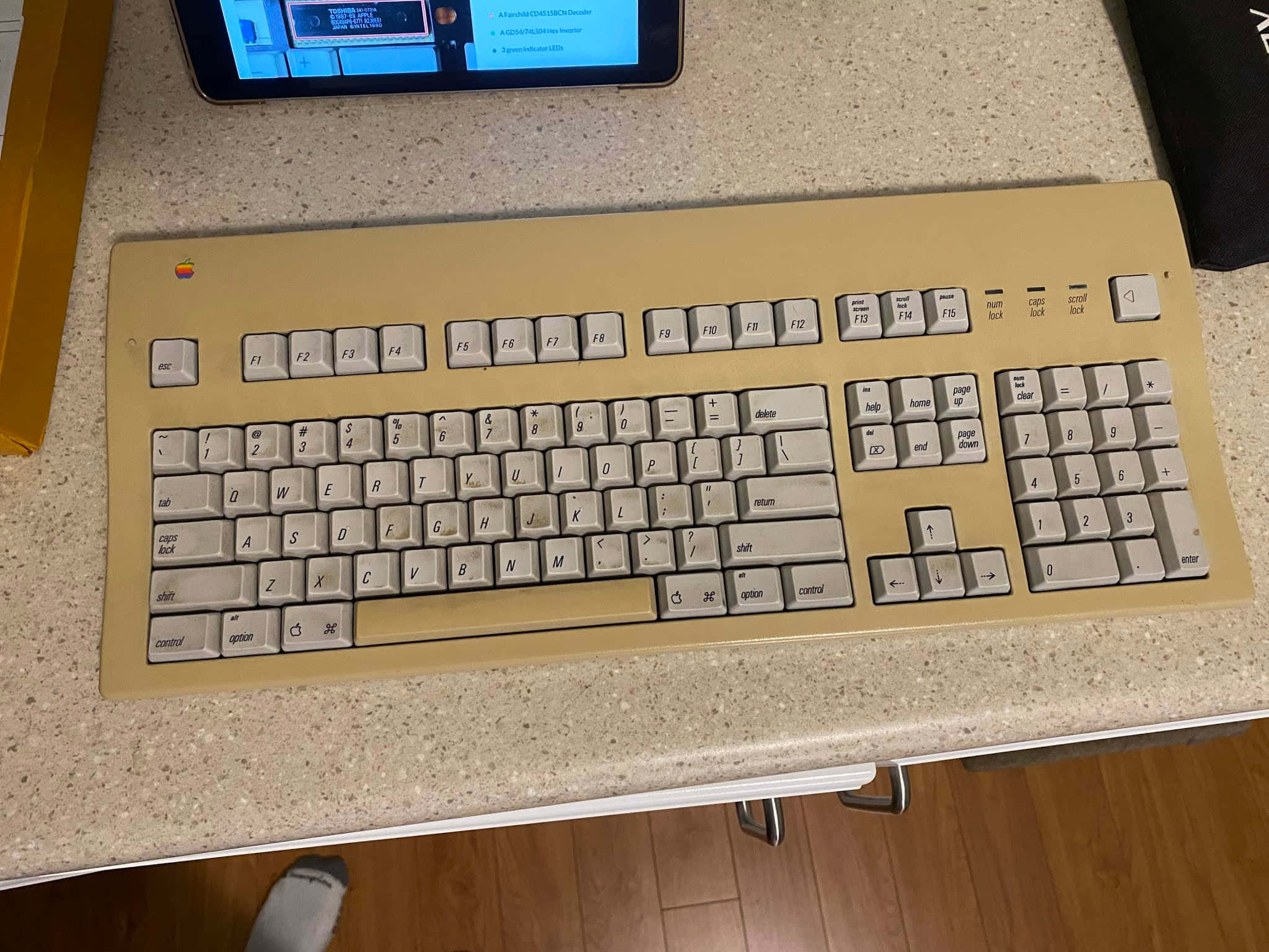 Retro Tech 桌布 壁紙 — 一個米色復古Apple鍵盤（白色按鍵）放在有斑點的檯面上，旁邊是一部顯示藍色螢幕的平板電腦。