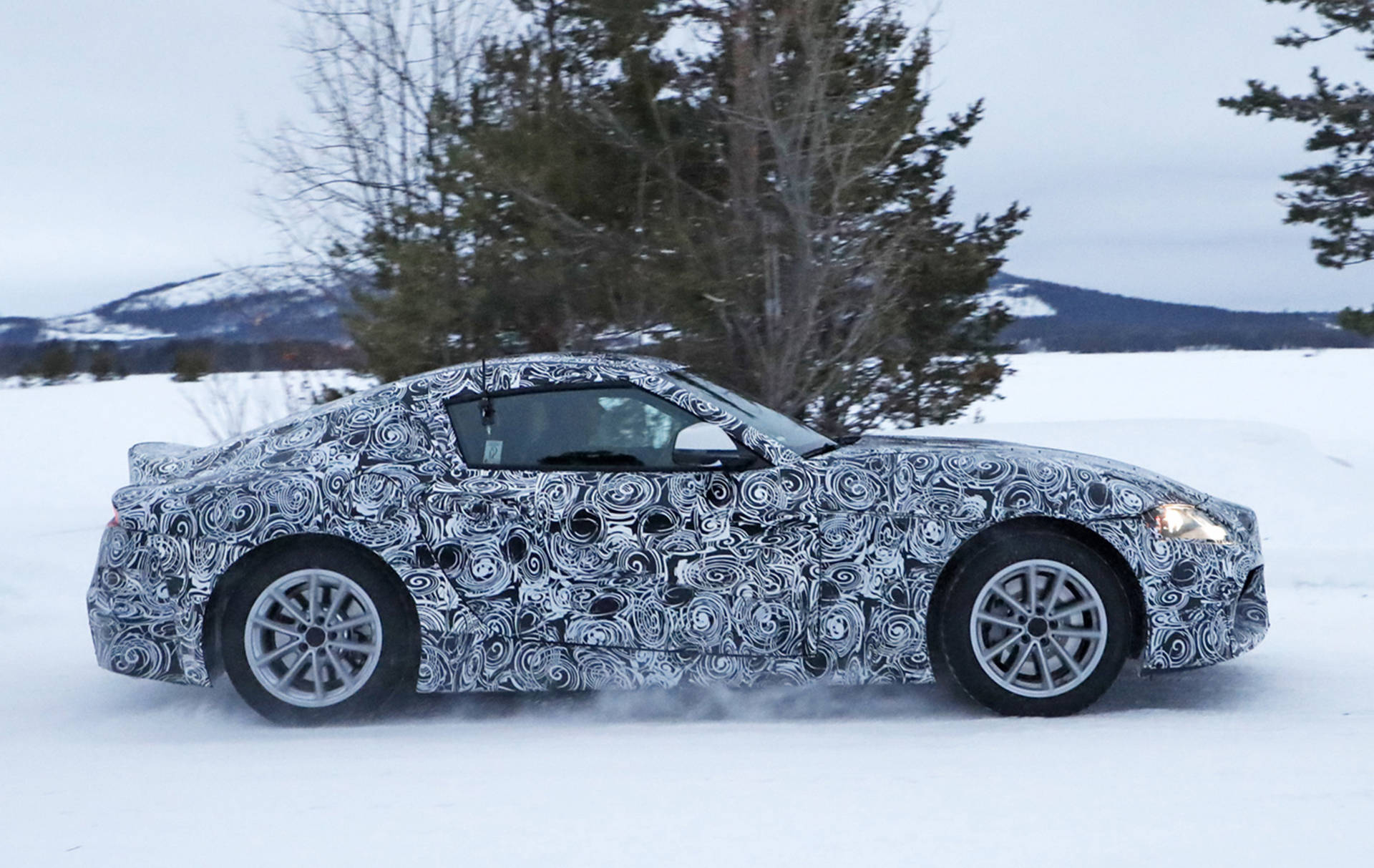 迷彩跑車在雪景中行駛，有樹木和山脈。 — Toyota Supra 壁紙