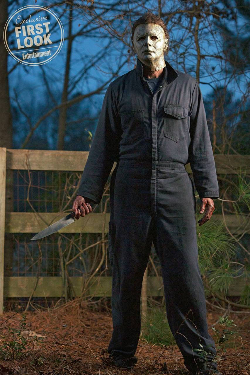 michael 桌布 壁紙 — Michael Myers 在黃昏時分站在樹林地區，手持刀，戴著標誌性的白色面具和深色連身衣，氣勢洶洶。