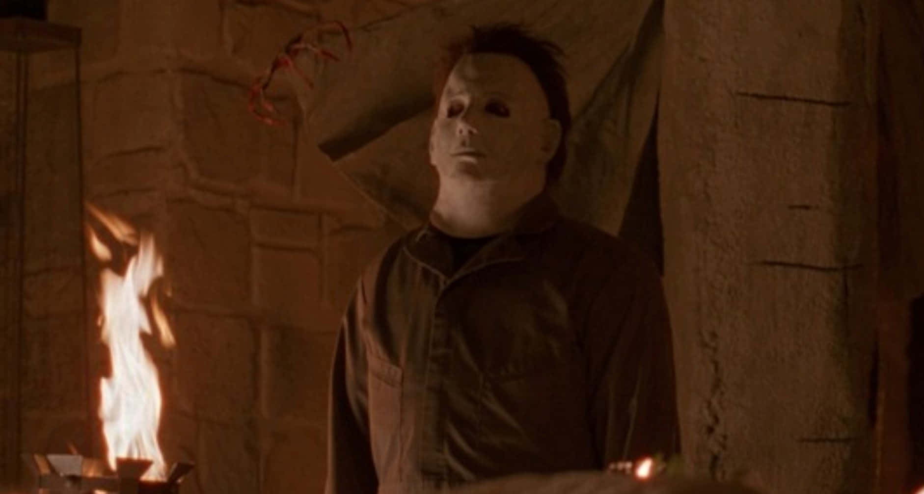 麥可·邁爾斯 桌布 壁紙 — Michael Myers 靜靜地站在昏暗的石室中，搖曳的火焰在牆上投射出詭異的影子。