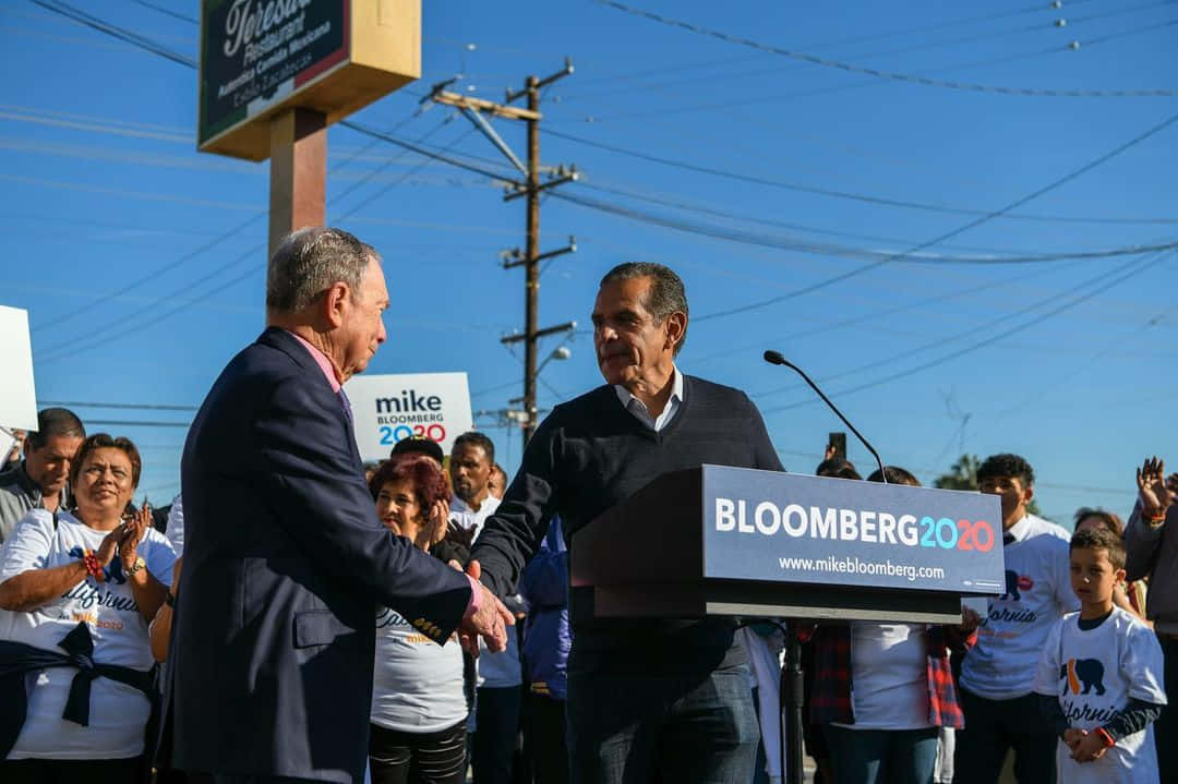 政治 桌布 壁紙 — Mike Bloomberg 在陽光明媚的戶外競選集會上與支持者握手，頭頂是藍天和電線。